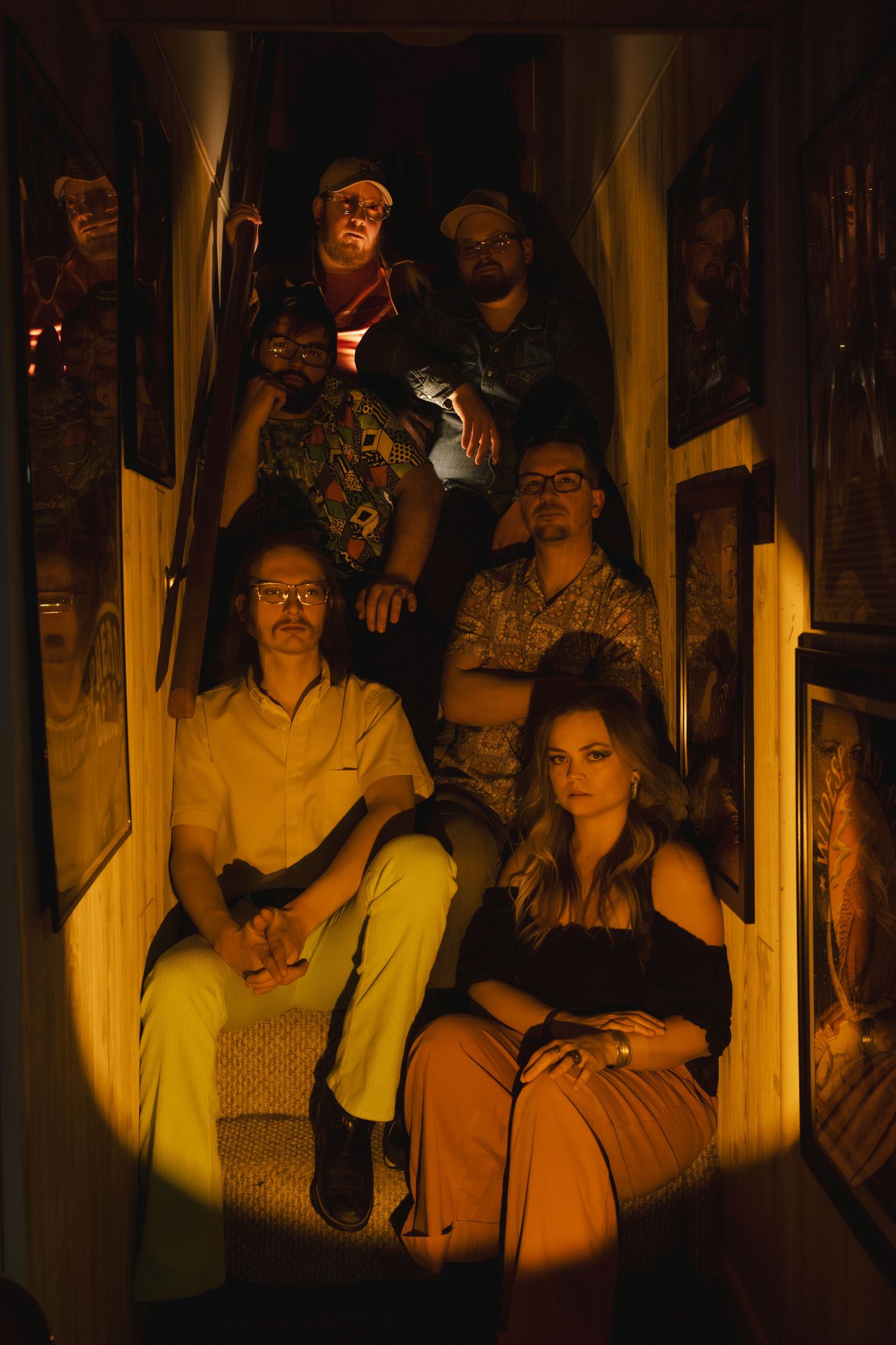 A group of people sit together on wooden stairs in dramatic warm lighting creating a moody atmospheric scene.