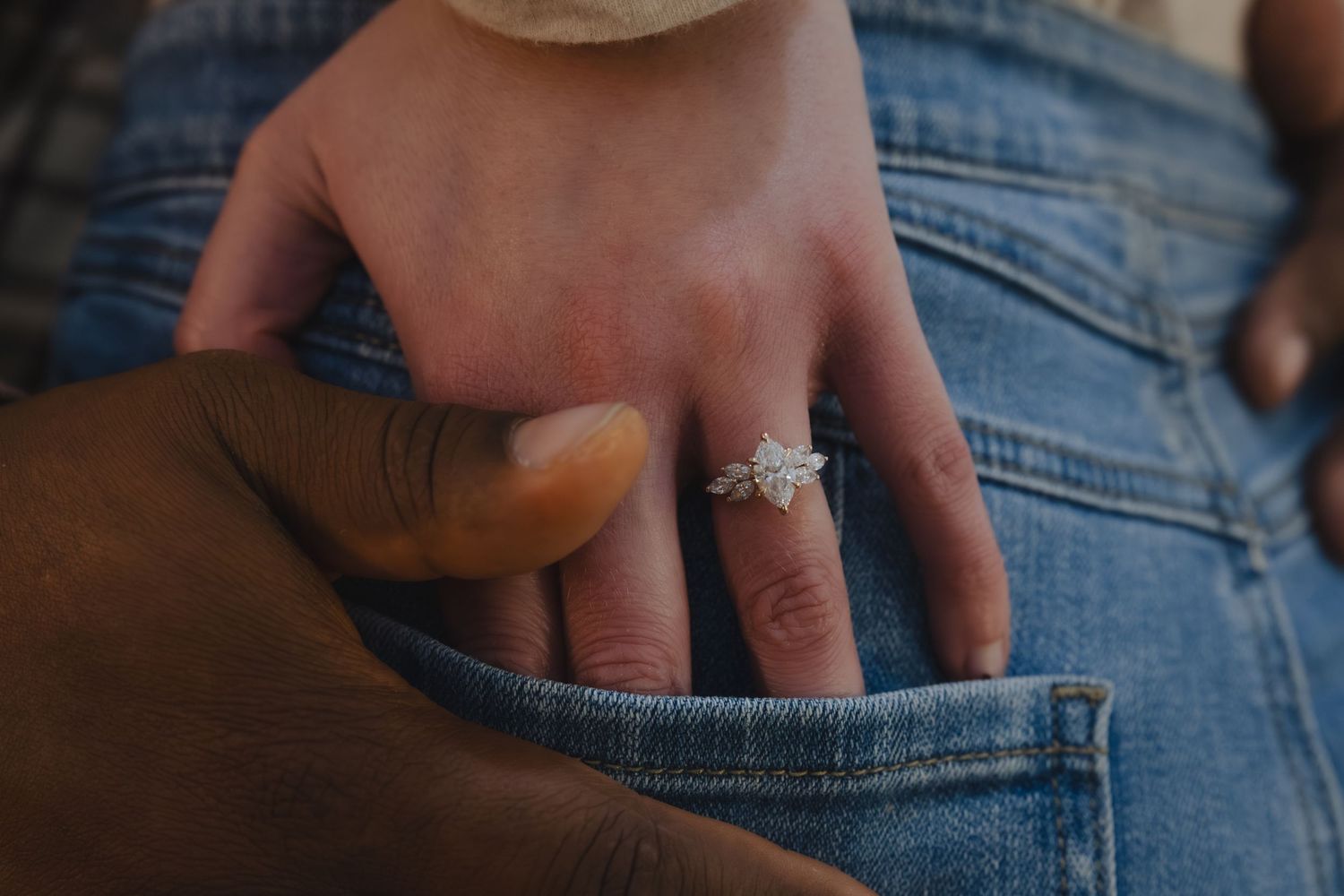 Close-up of a hand wearing a diamond engagement ring resting on a denim pocket.
