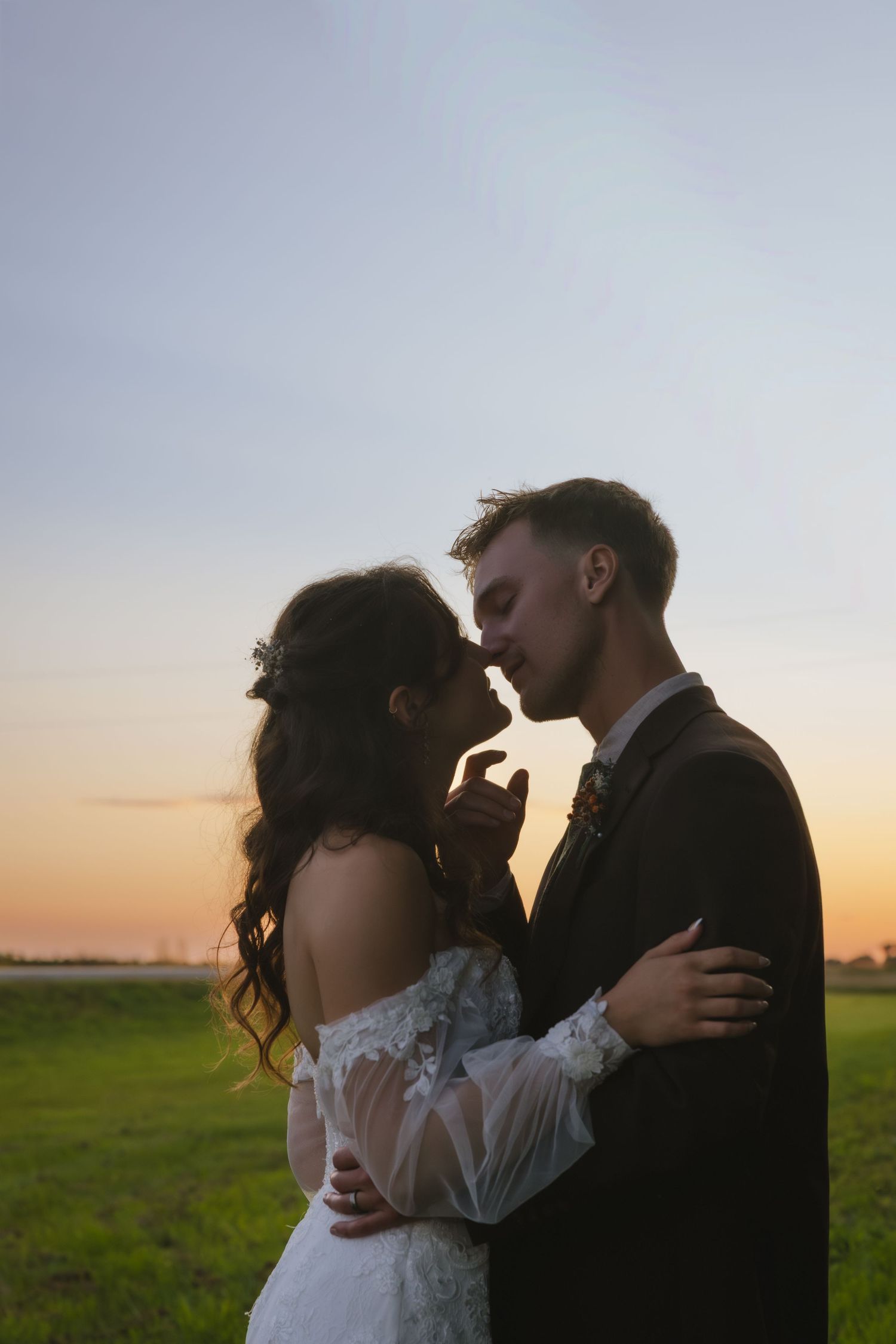 A couple in formal wedding attire embraces and kisses in a field at sunset, creating a romantic silhouette.