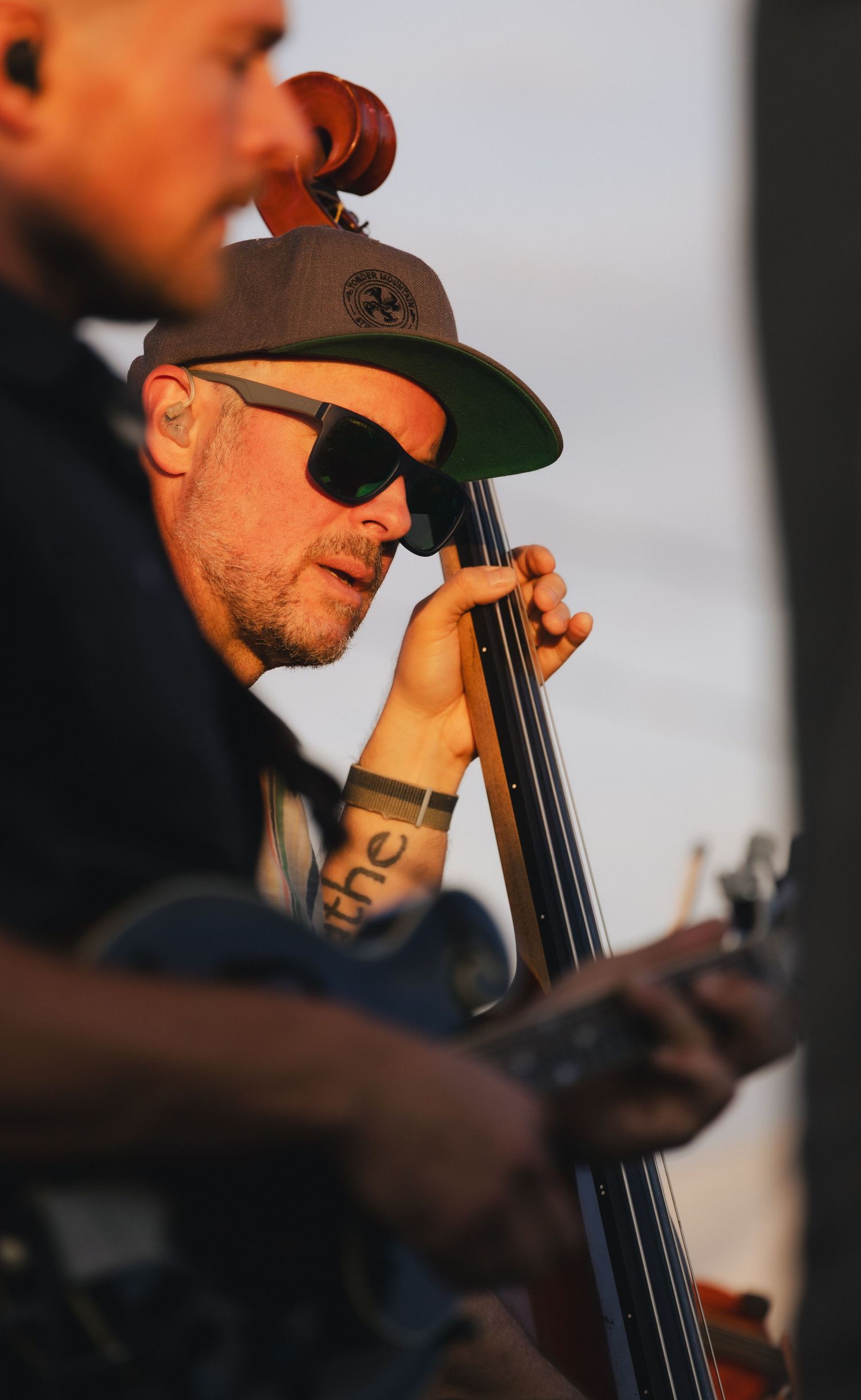 Tattooed musician playing double bass outdoors, wearing sunglasses and a gray cap, captured in warm golden hour light.