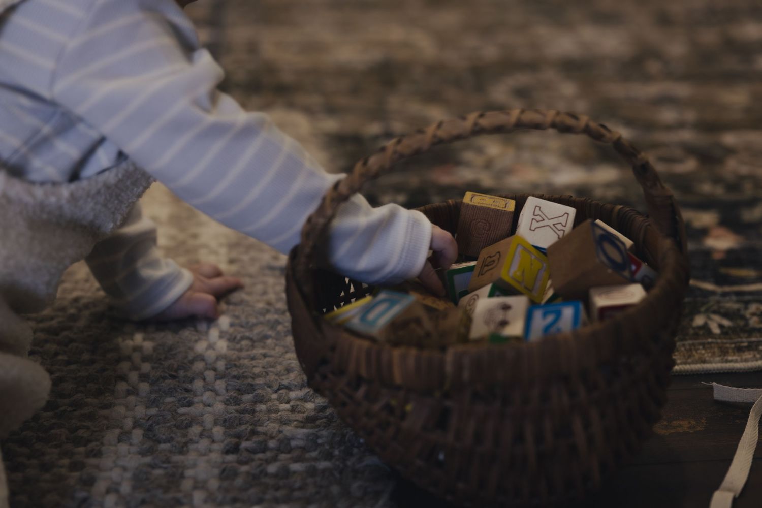 Child reaching into a woven basket filled with colorful small toy blocks on a patterned rug.