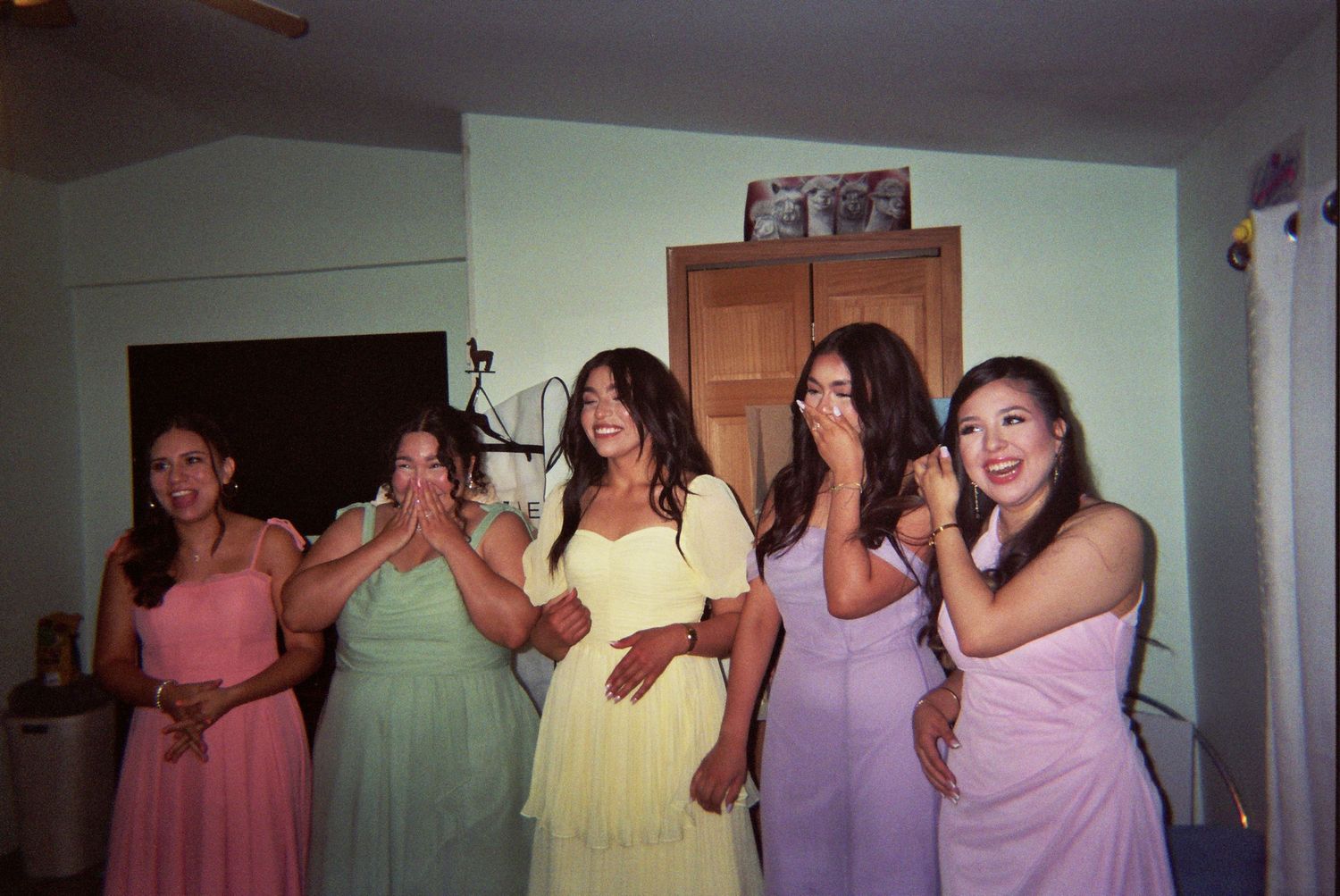 Five friends in pastel colored dresses stand together in a row making expressive gestures at a party.