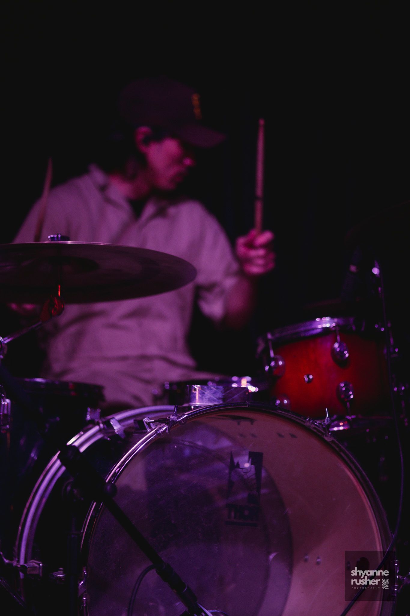 Drummer playing behind a drum kit bathed in purple stage lighting with drum kick in foreground.