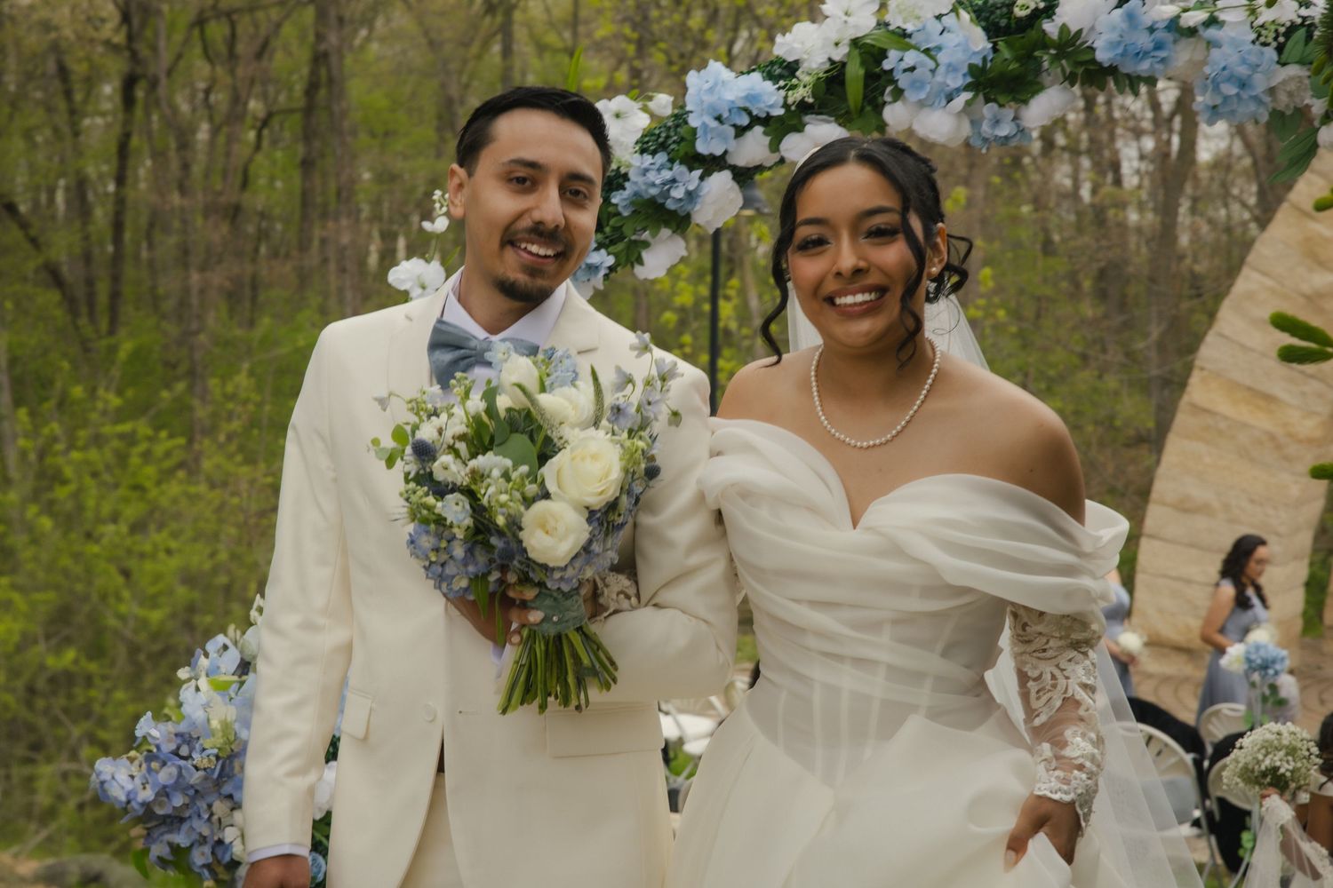 A happy couple on their wedding day wearing white attire and holding a bouquet with blue and white flowers in a wooded setting.