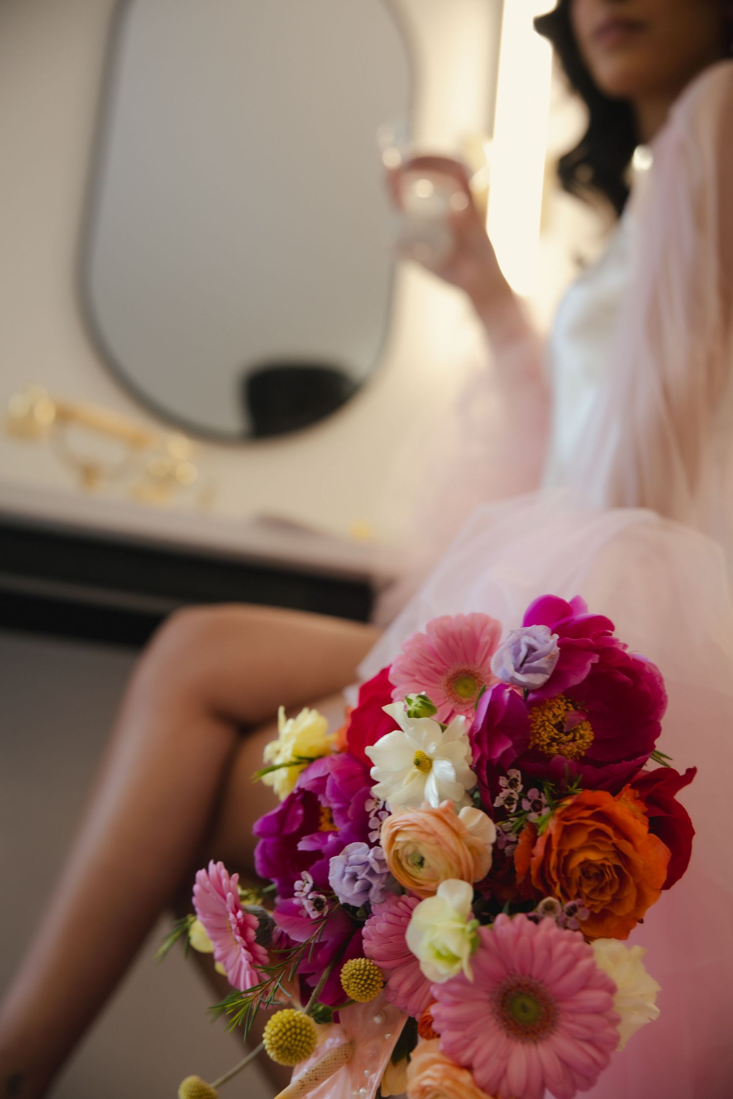 Vibrant floral bouquet with pink gerbera daisies, orange roses and purple blooms near vanity mirror in soft lighting.