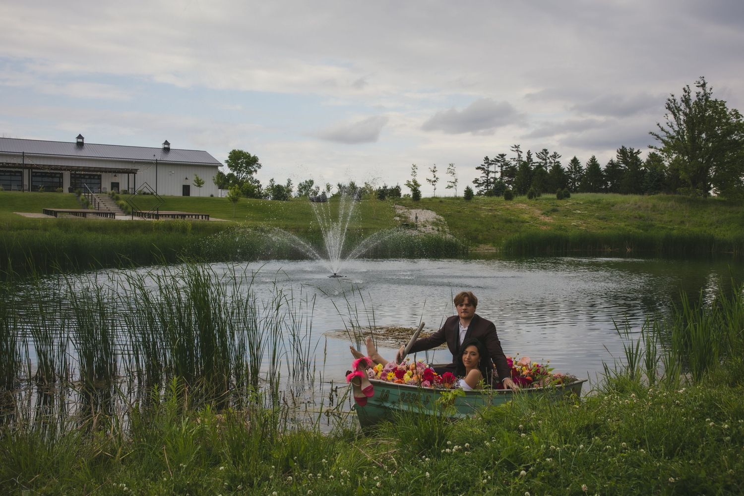 A small fountain sprays in a pond while someone sits at the grassy bank with picnic items.