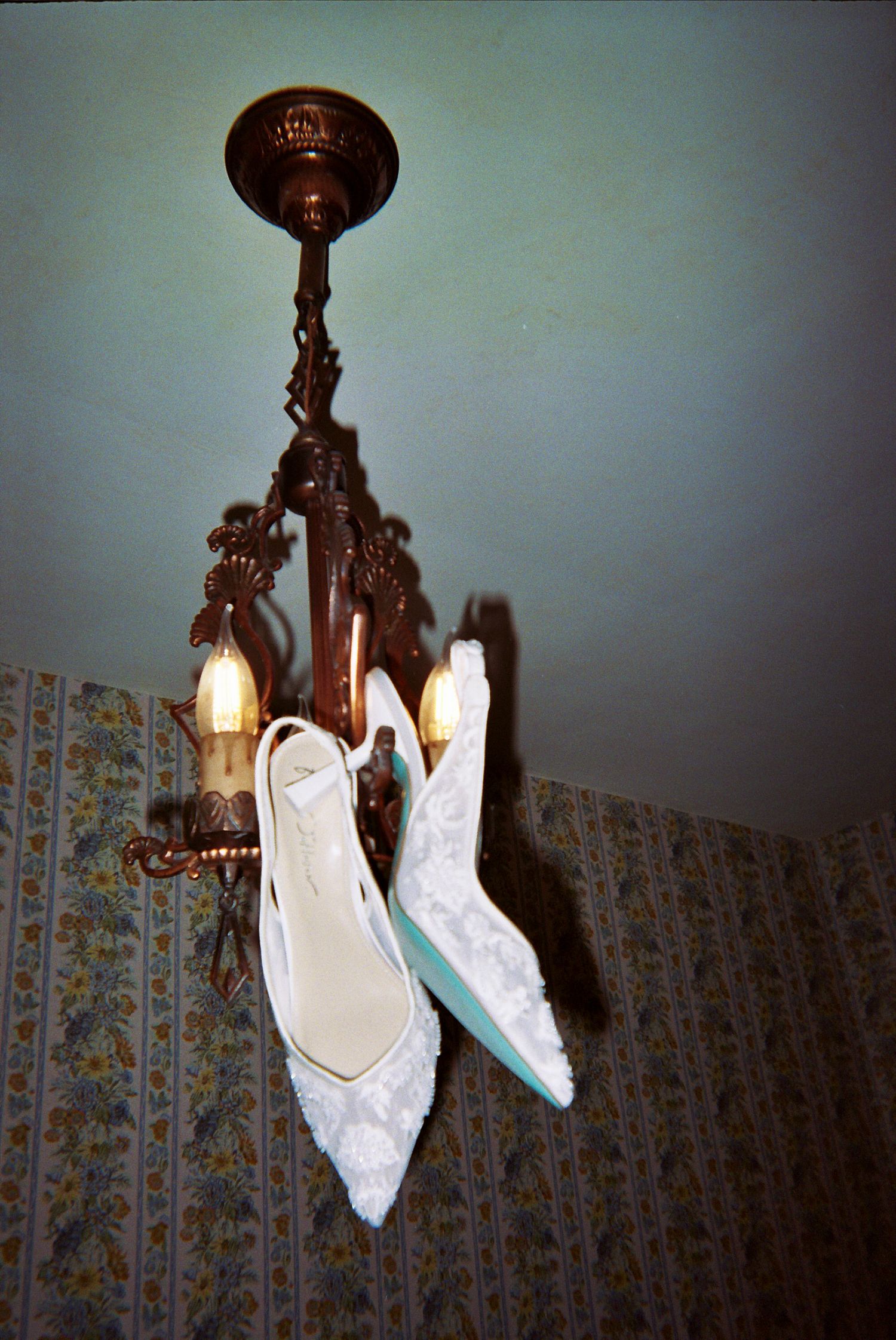 White lace wedding shoes hanging from an ornate vintage chandelier against floral wallpaper.
