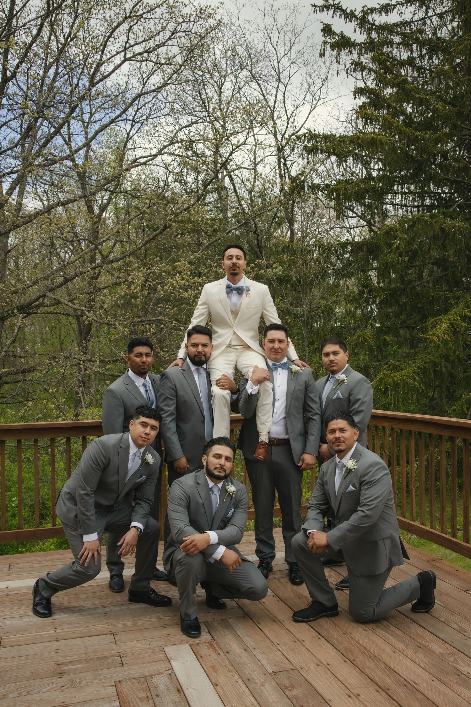 Wedding party of groomsmen in gray suits pose together on wooden deck with trees in background.