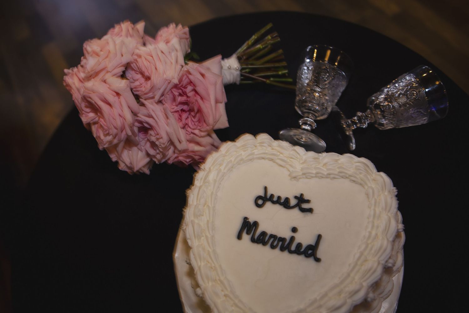A heart-shaped cake with 'Just Married' text sits on a black plate next to pink roses and crystal glasses.