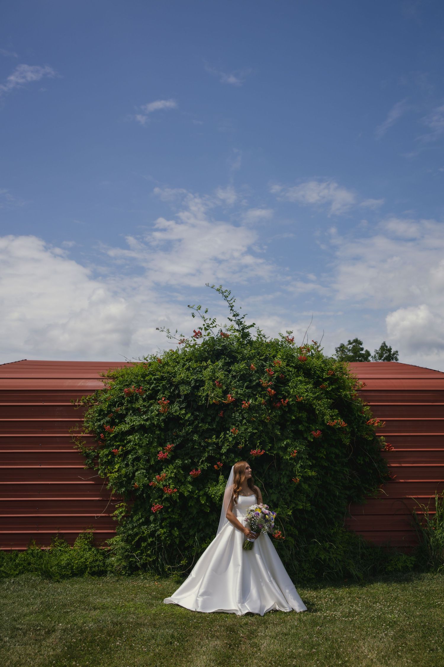 Bride in elegant white wedding gown poses beside flowering bush against rustic brown wall under blue sky.