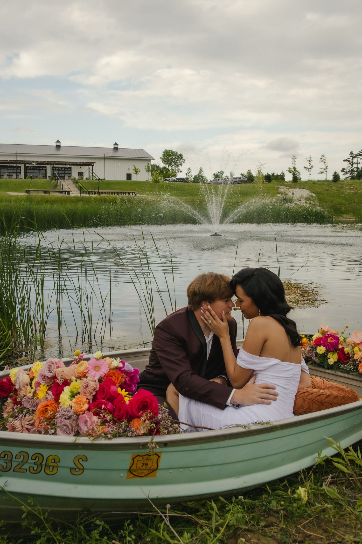 A romantic couple sits in a boat on a pond with vibrant flowers and a fountain spraying in the background.