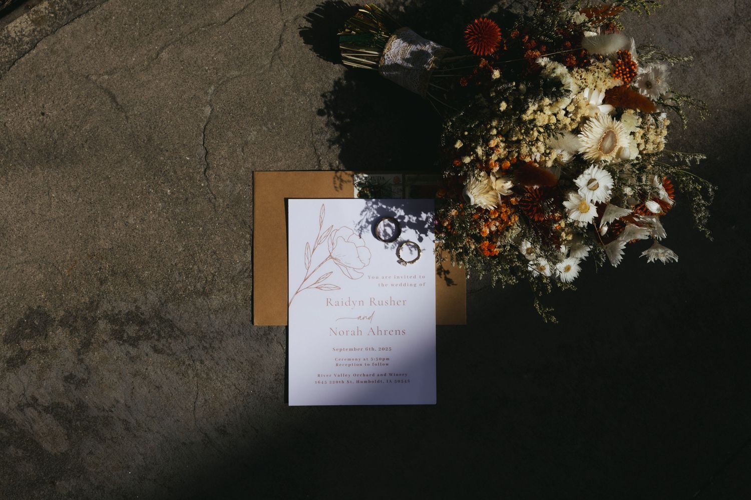 A stylish wedding invitation sits among delicate white and blush floral arrangements and greenery on a dark surface.