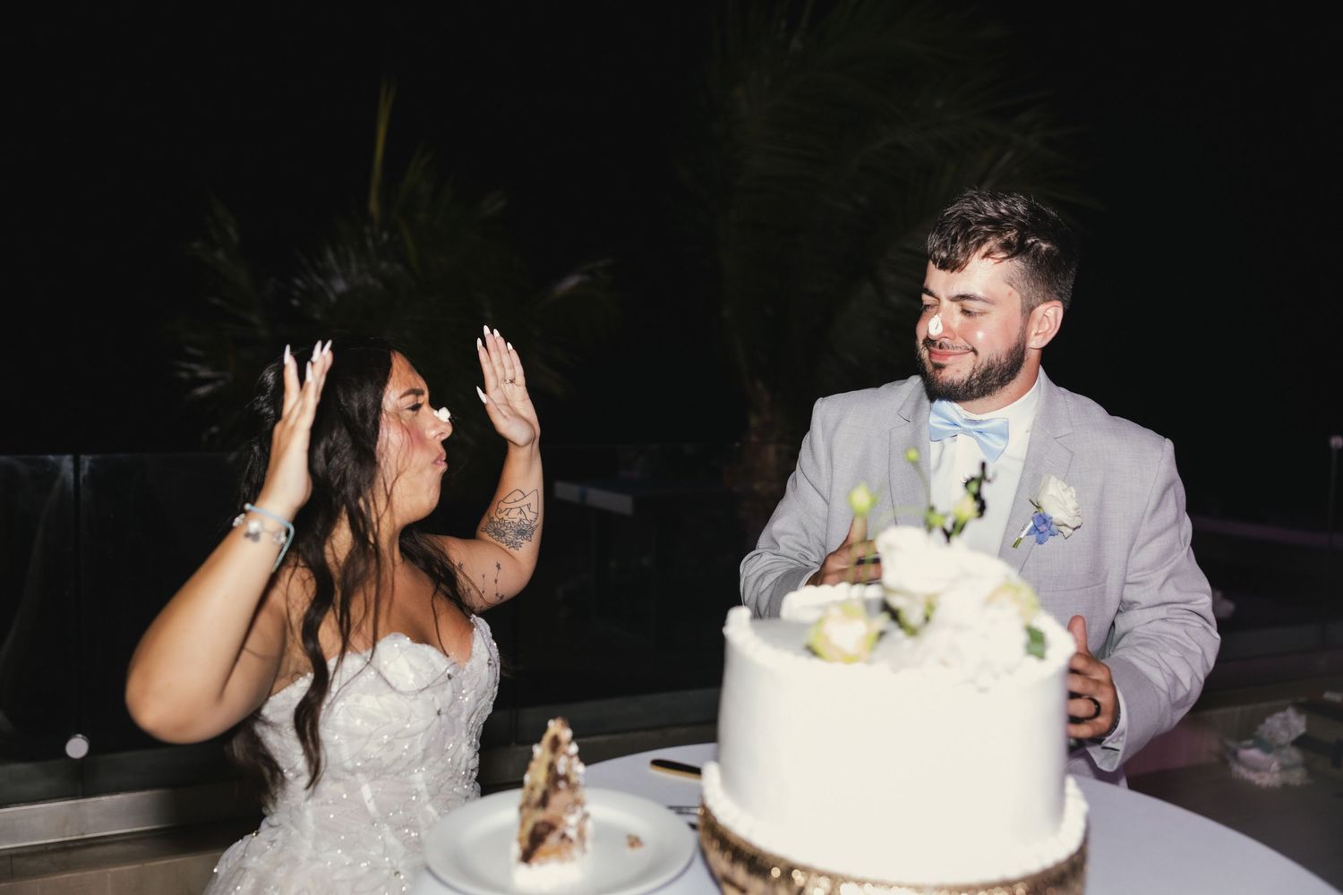 Bride and groom laughing while cutting their white wedding cake at an outdoor evening reception.