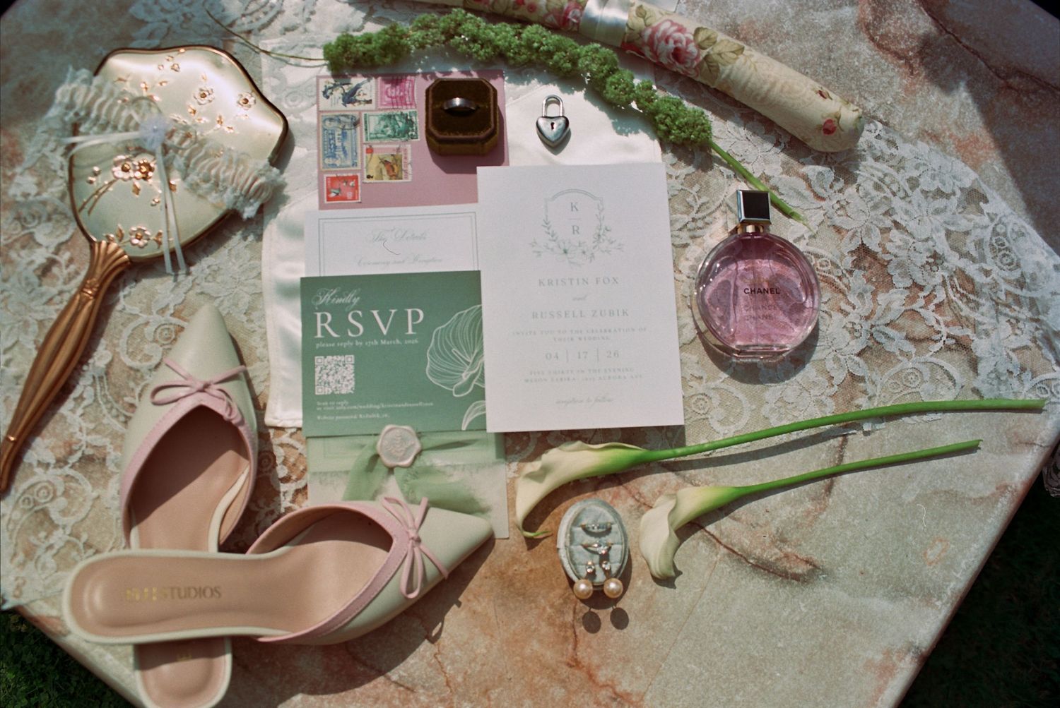 Elegant wedding flat lay with RSVP card, invitation, pink heels, perfume, ring box, and calla lilies on marble.