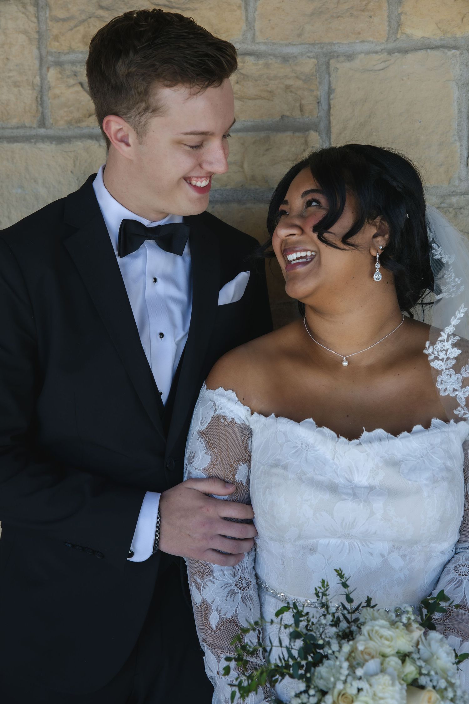 A joyful wedding couple shares a candid laughing moment together against a stone wall background.