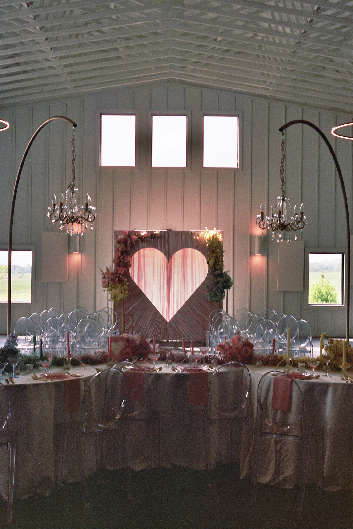 Elegant wedding reception setup with heart backdrop, crystal chandeliers and soft pink lighting in a white barn venue.