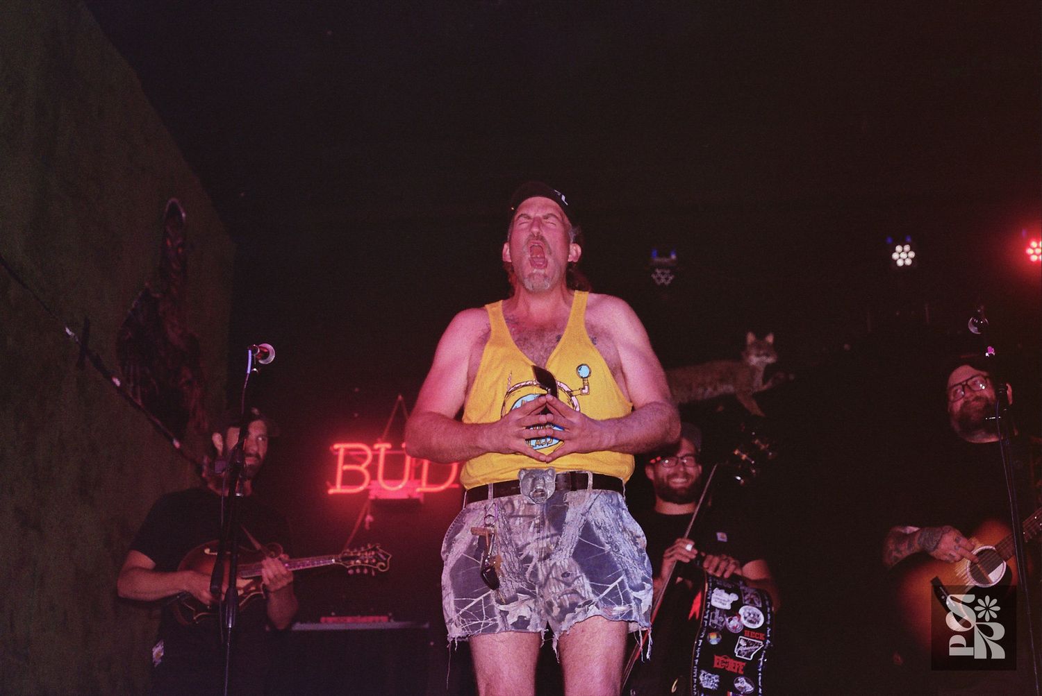 Musical performer in yellow tank top and patterned shorts on stage with red neon lighting in background.
