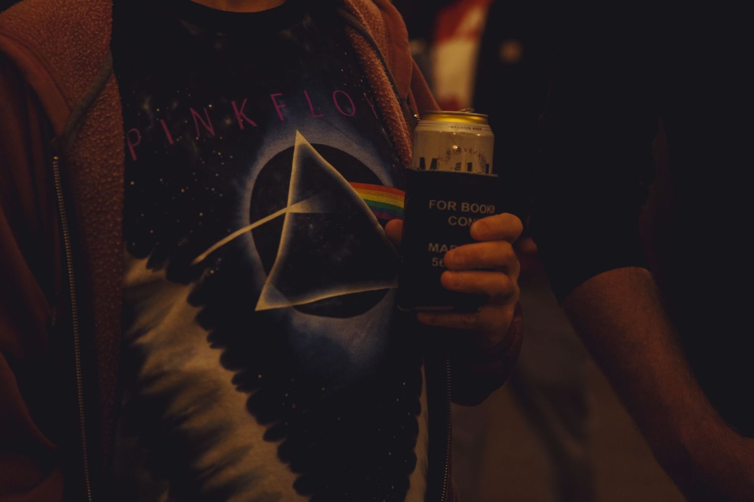 Someone wearing a Pink Floyd Dark Side of the Moon t-shirt holds a drink in a dimly lit setting.