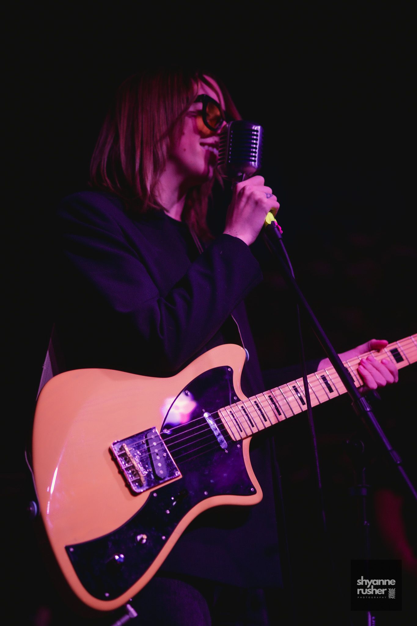Musician performs on stage holding a tan electric guitar under dramatic purple lighting at a concert.