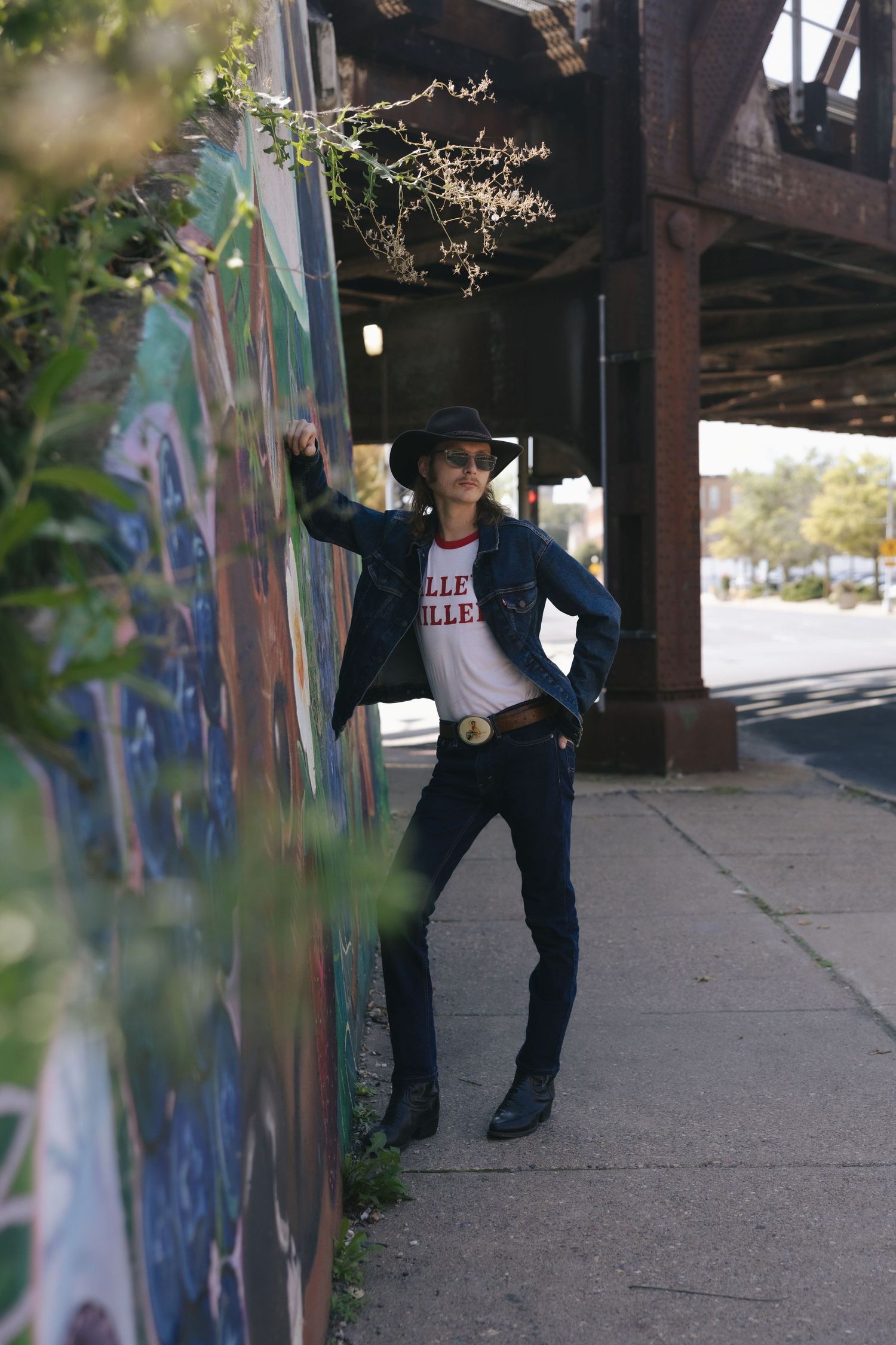 A stylish man in a cowboy hat, denim jacket, and boots leans against a colorful mural on an urban sidewalk.