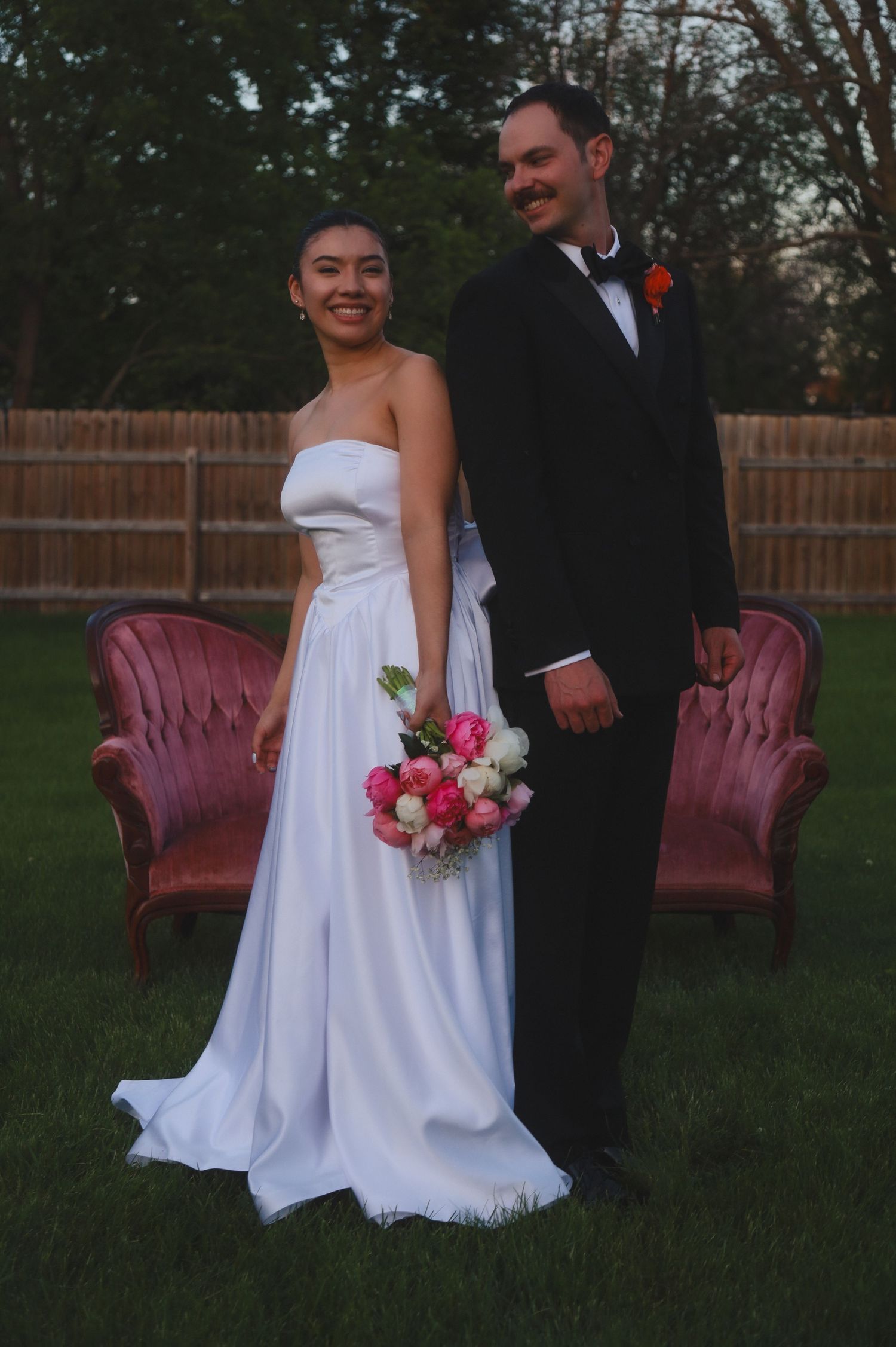A couple in formal wedding attire poses in a backyard with pink roses and vintage velvet chairs.