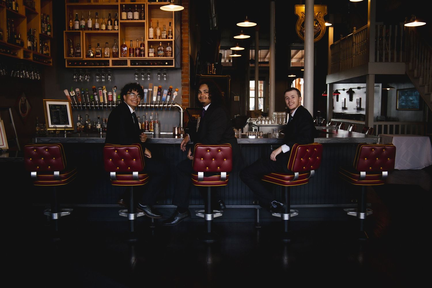 Three people in formal attire sit at a dimly lit vintage bar with red leather barstools in a classic establishment.