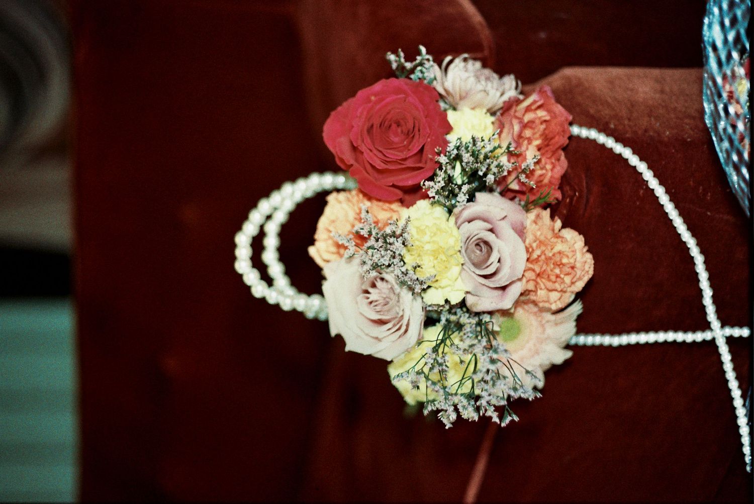 Elegant pearl necklace draped next to colorful floral arrangements with roses and baby's breath on dark background.