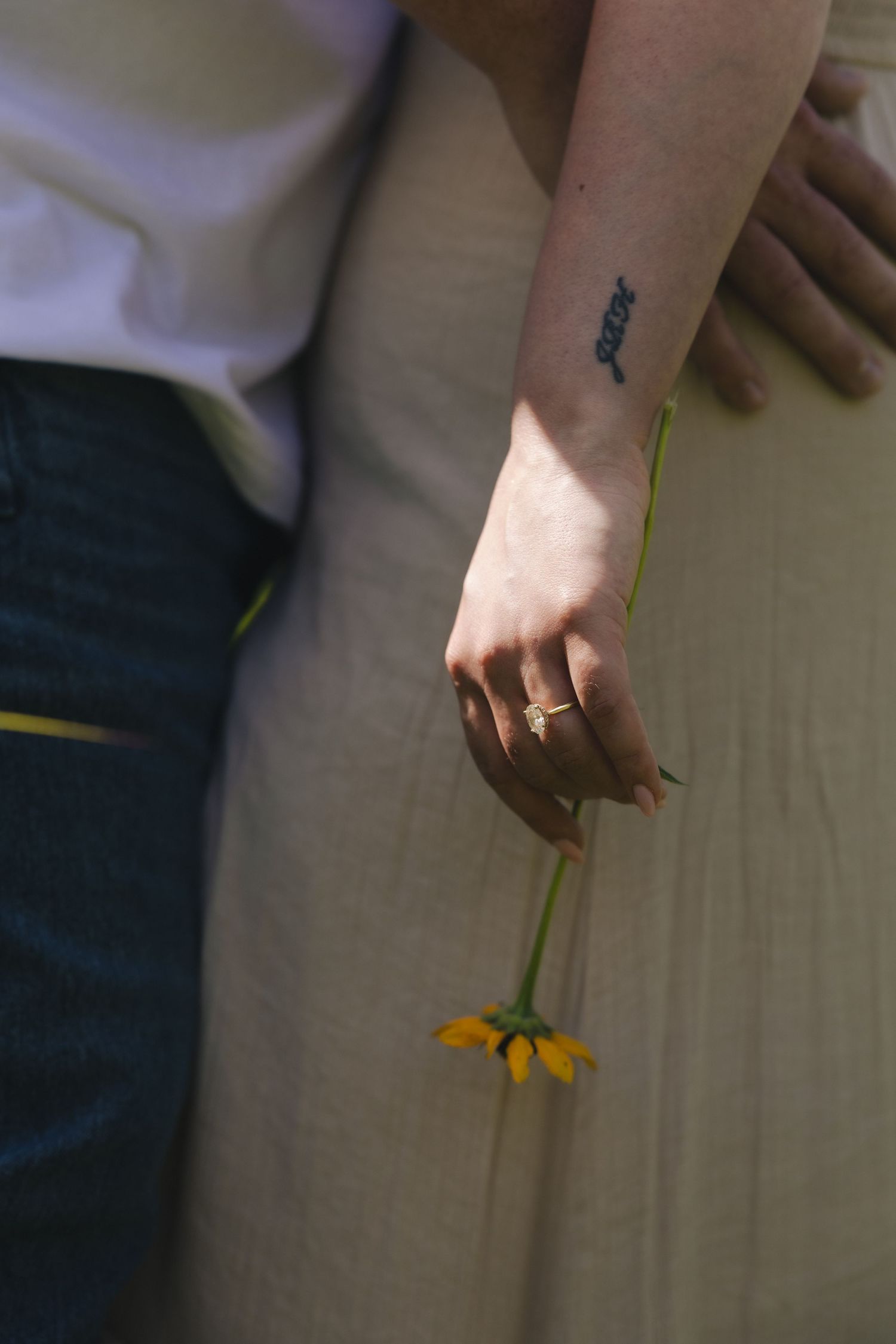 A woman's hand holding a yellow daisy flower, wearing a gold ring, with a small tattoo on her wrist.