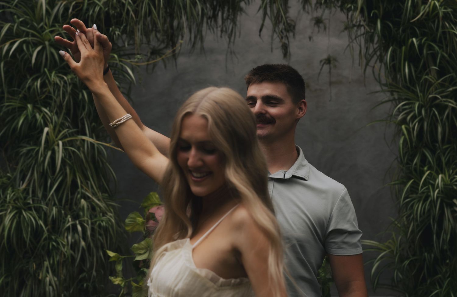 Couple poses romantically among lush green tropical foliage, woman in white dress raises arm gracefully.