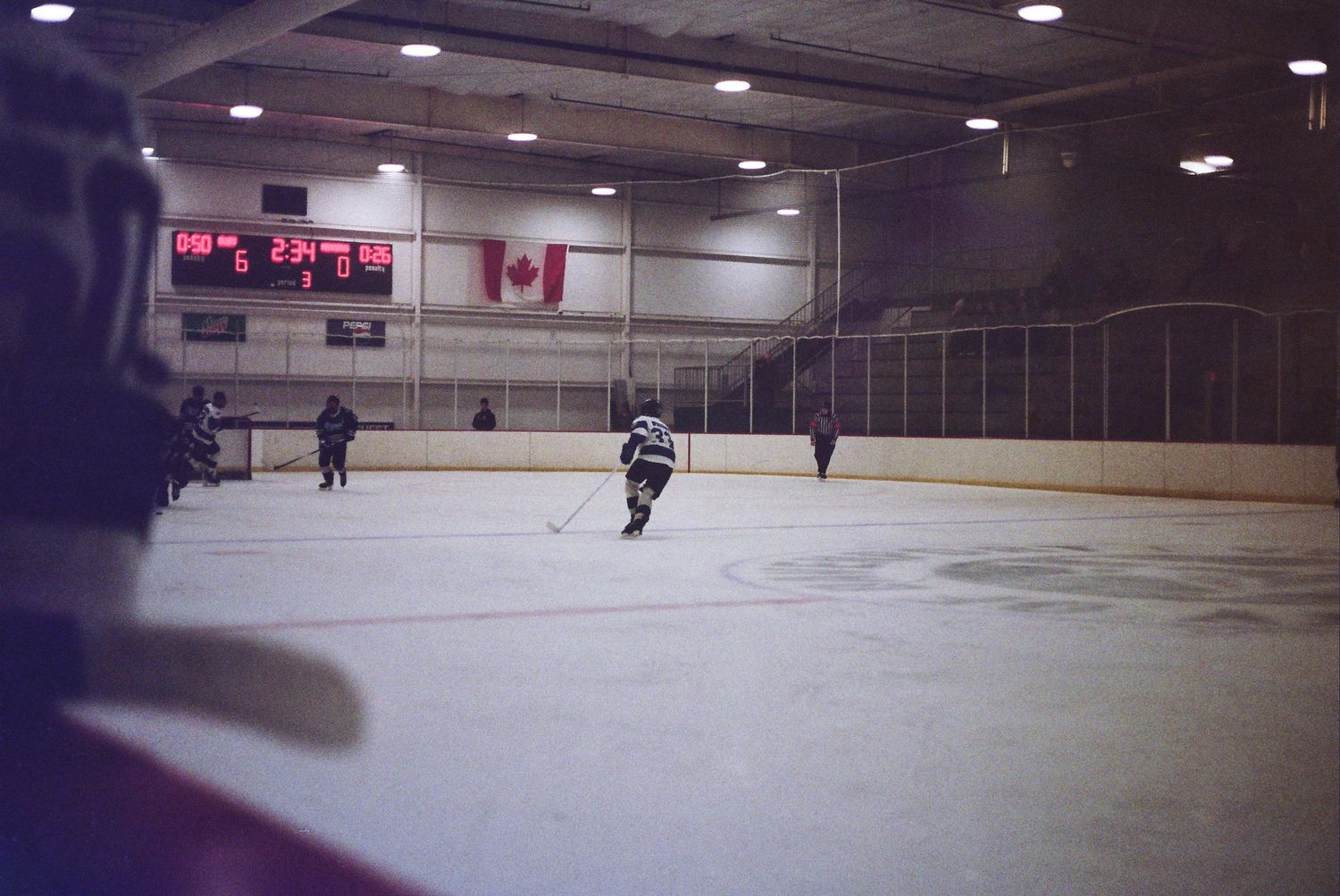 Hockey players skating on an indoor ice rink with a Canadian flag displayed on the arena wall.
