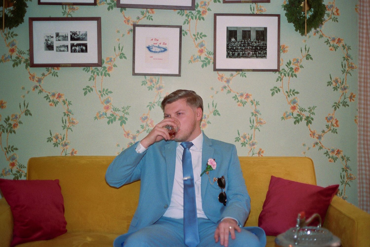 Man in light blue suit sitting on yellow sofa, drinking, with floral wallpaper and framed photos behind him.