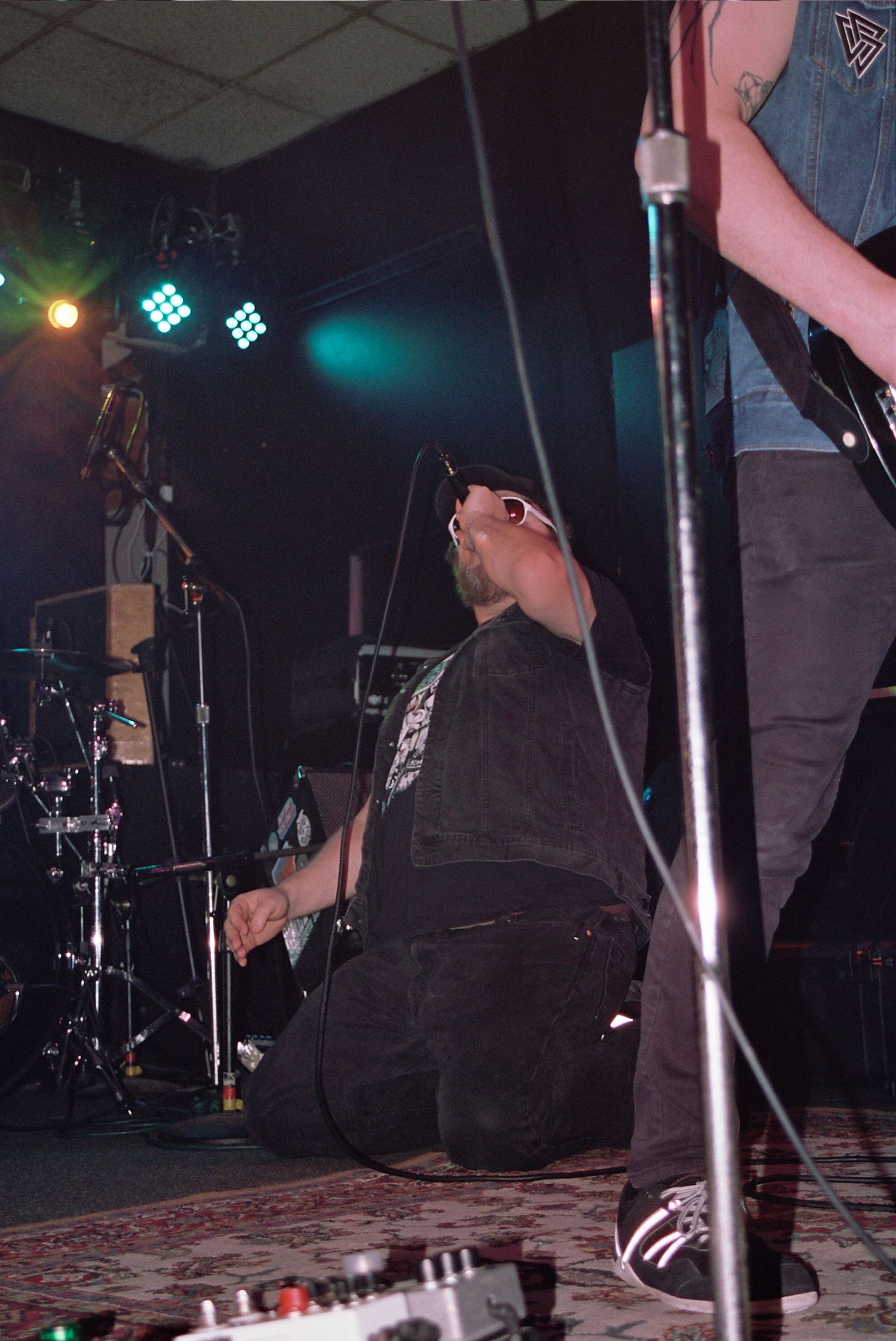 Musician kneeling on stage during intense rock concert performance with dramatic lighting and band equipment visible.