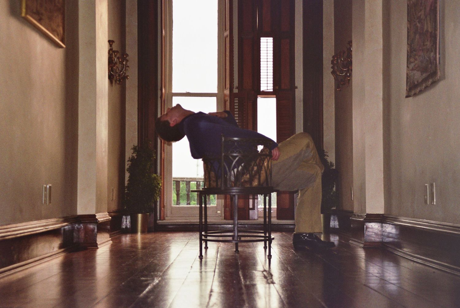Person bending over a chair in an elegant hallway with hardwood floors and tall windows.