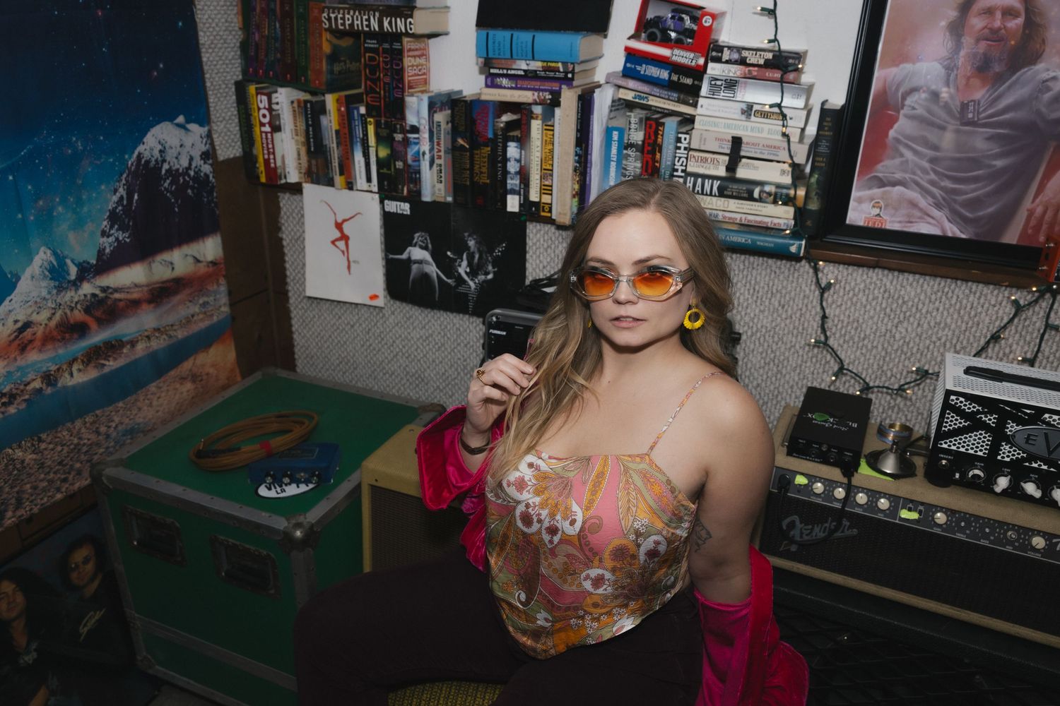Young woman in colorful top and orange sunglasses poses in a retro room filled with books and vinyl records.