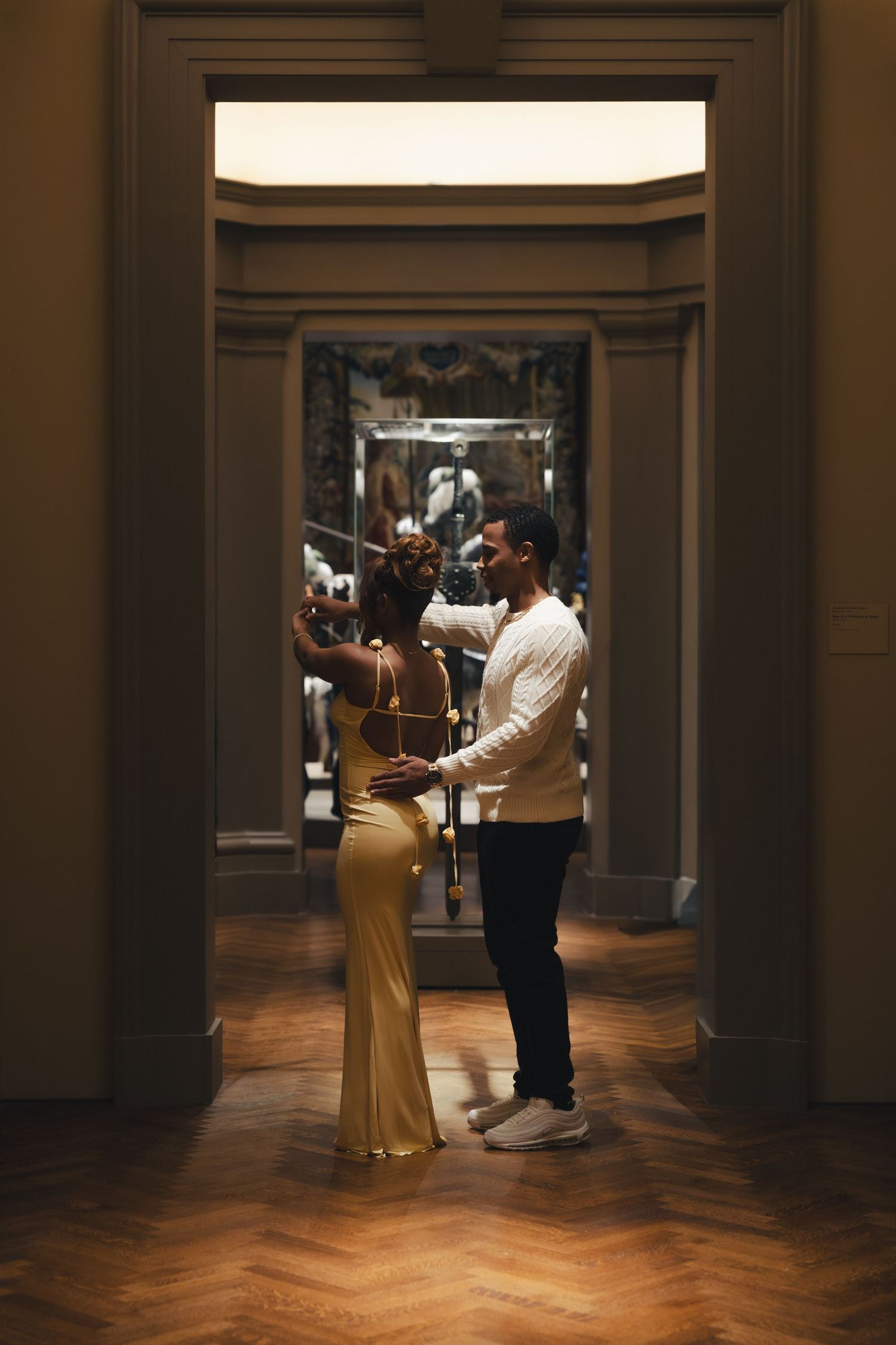 Couple dancing elegantly in a grand museum hallway, woman in gold gown, man in white sweater and black pants.