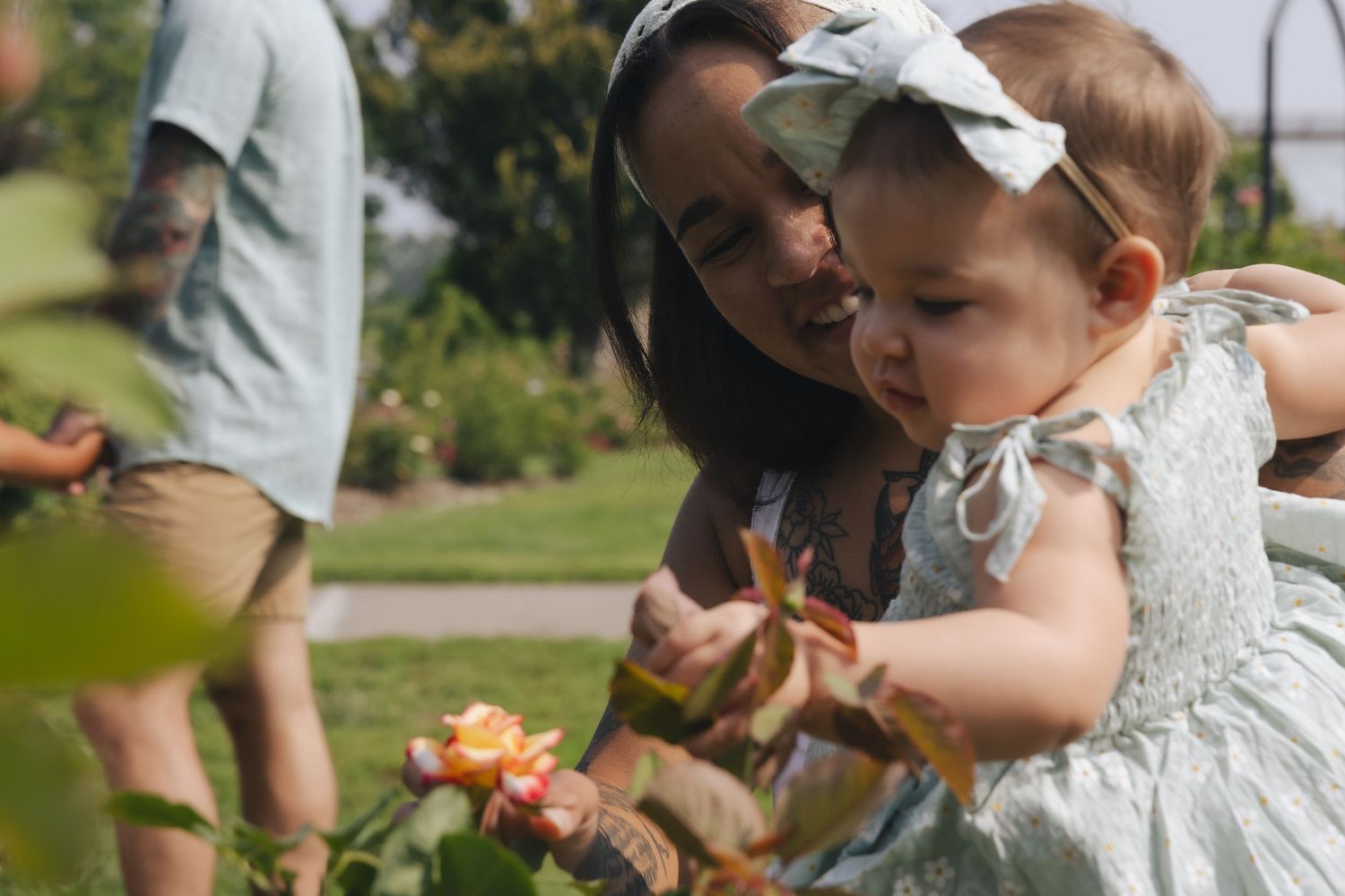 A mother holds her baby girl near blooming roses in a sunny garden, both wearing light dresses.