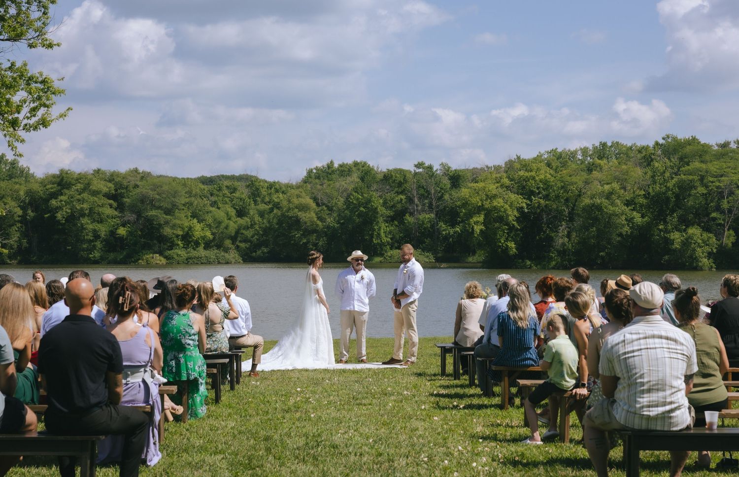 Outdoor wedding ceremony by a lake with guests seated on benches, couple at altar, lush green trees in background.