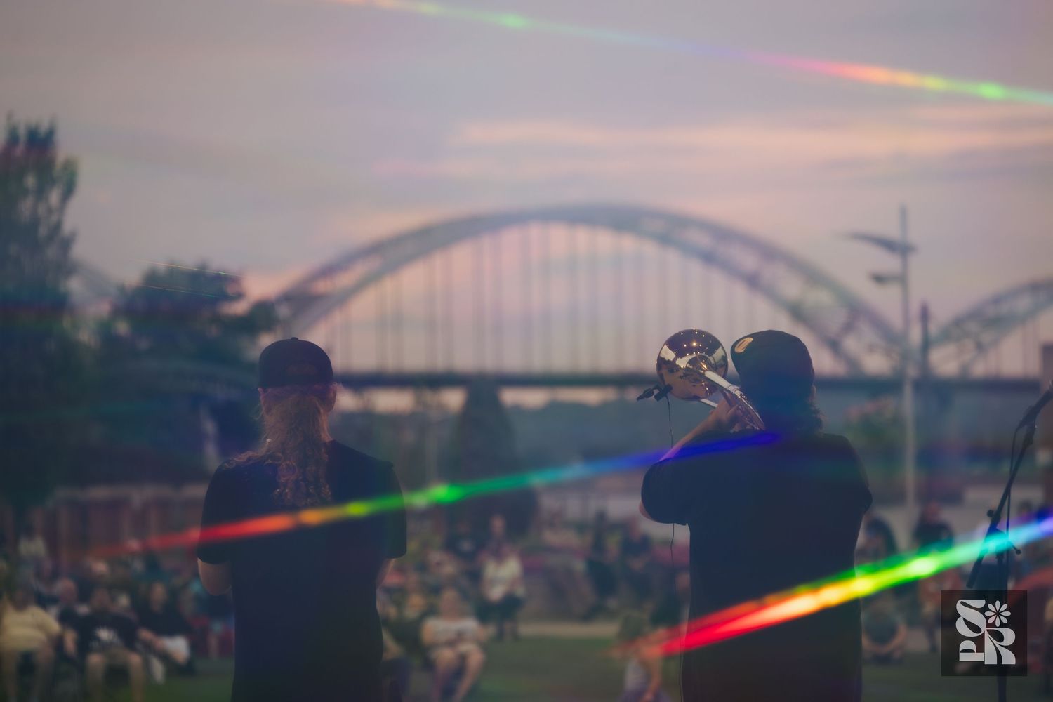 Musician plays trumpet at outdoor concert near arched bridge at dusk with rainbow lens flare effect.