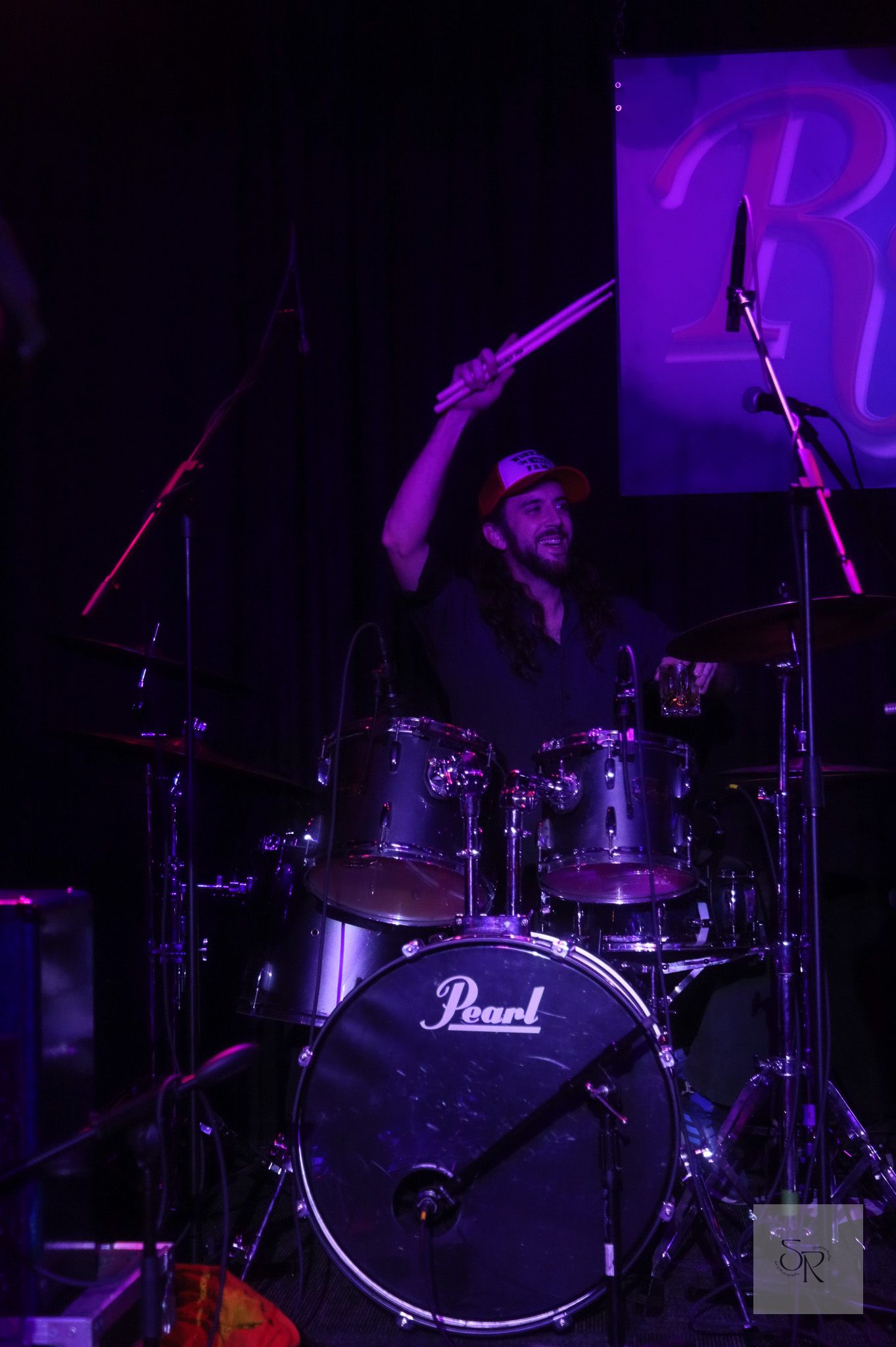 Drummer performs on a Pearl drum kit on stage illuminated by purple lighting during a live concert performance.