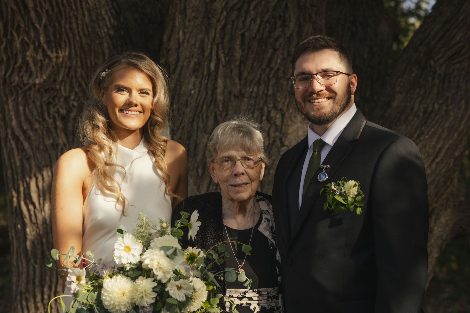 A wedding party poses together in a garden setting with trees and greenery in the background.