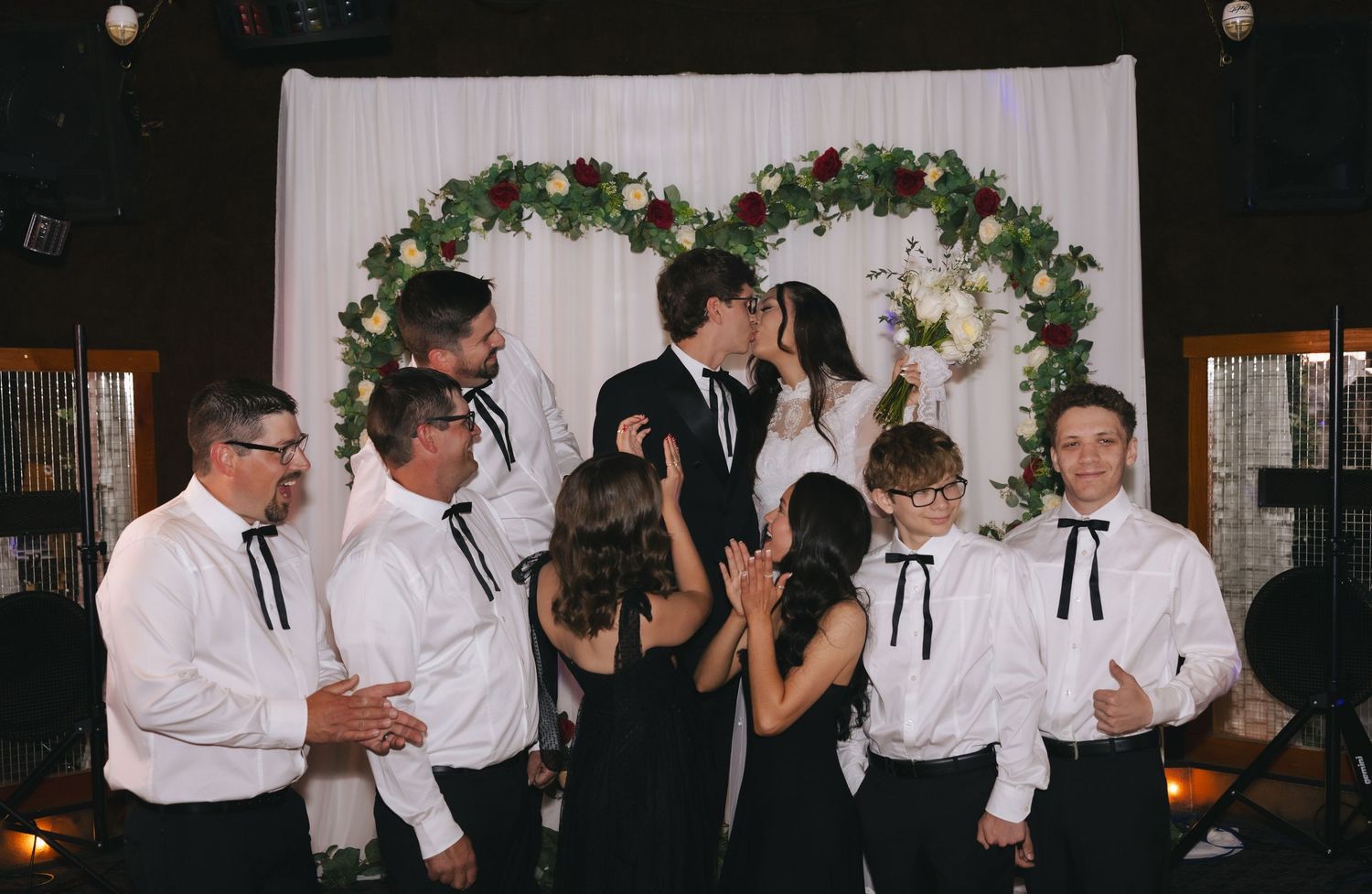 Wedding couple kissing at floral arch backdrop surrounded by bridal party in white shirts with black bow ties.