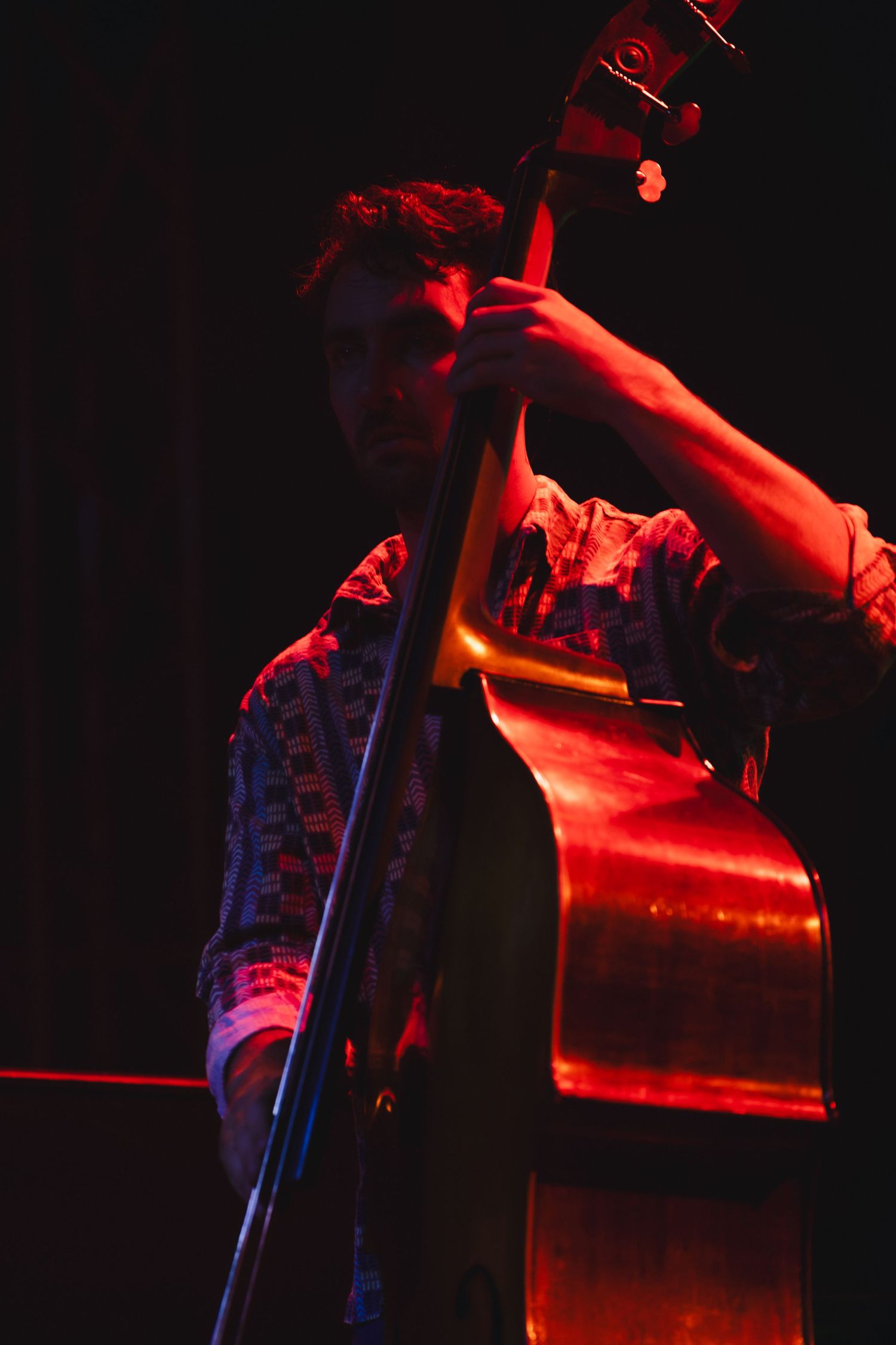 Musician playing upright double bass on stage under dramatic red lighting in a dark venue.