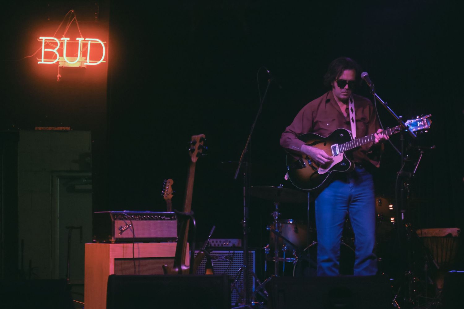 A musician performs on stage with an electric guitar under a red neon Bud sign in a dimly lit venue.