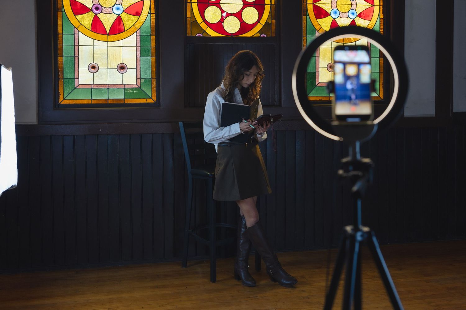 A figure stands in front of colorful stained glass windows while being filmed with a camera on a tripod.
