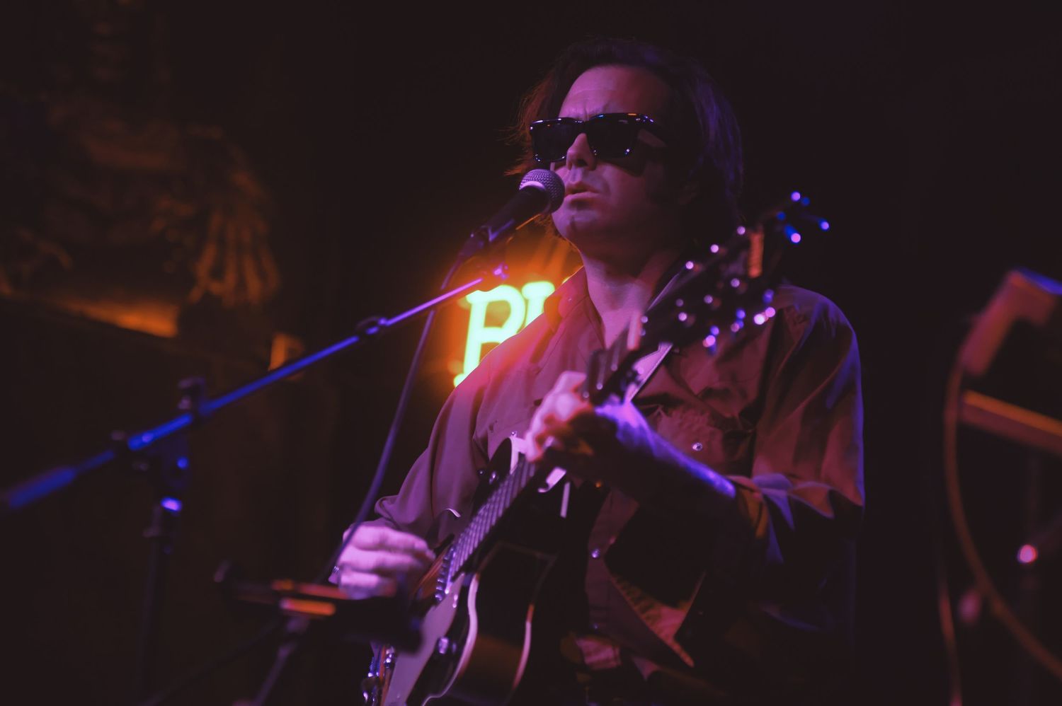 Musicians perform on stage in a dark venue with purple lighting creating a moody concert atmosphere.