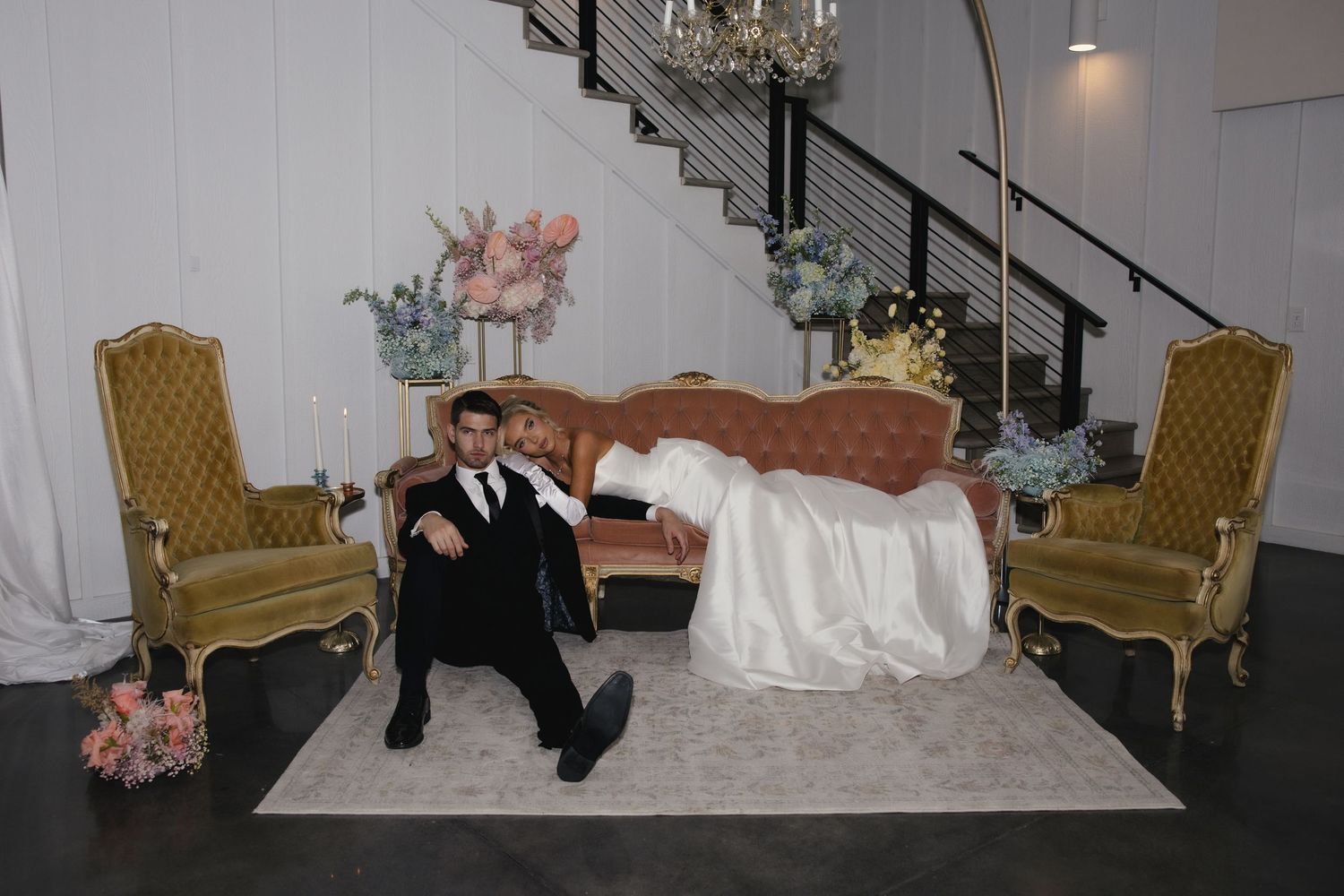 Newlyweds pose playfully on vintage furniture with gold chairs and floral decorations in an indoor venue setting.