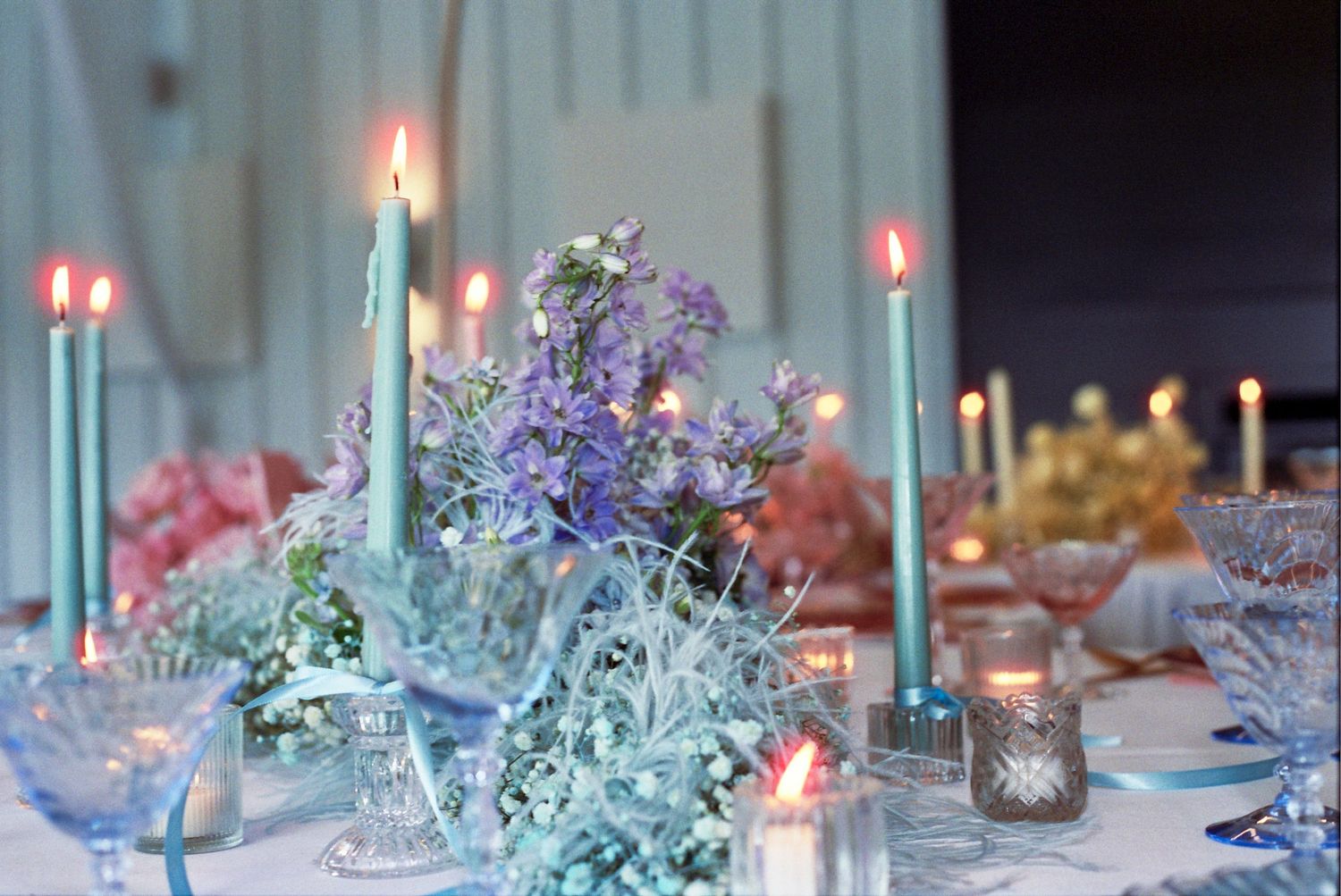 Elegant table setting with lit mint green candles and purple flowers surrounded by delicate glassware and ethereal decor.