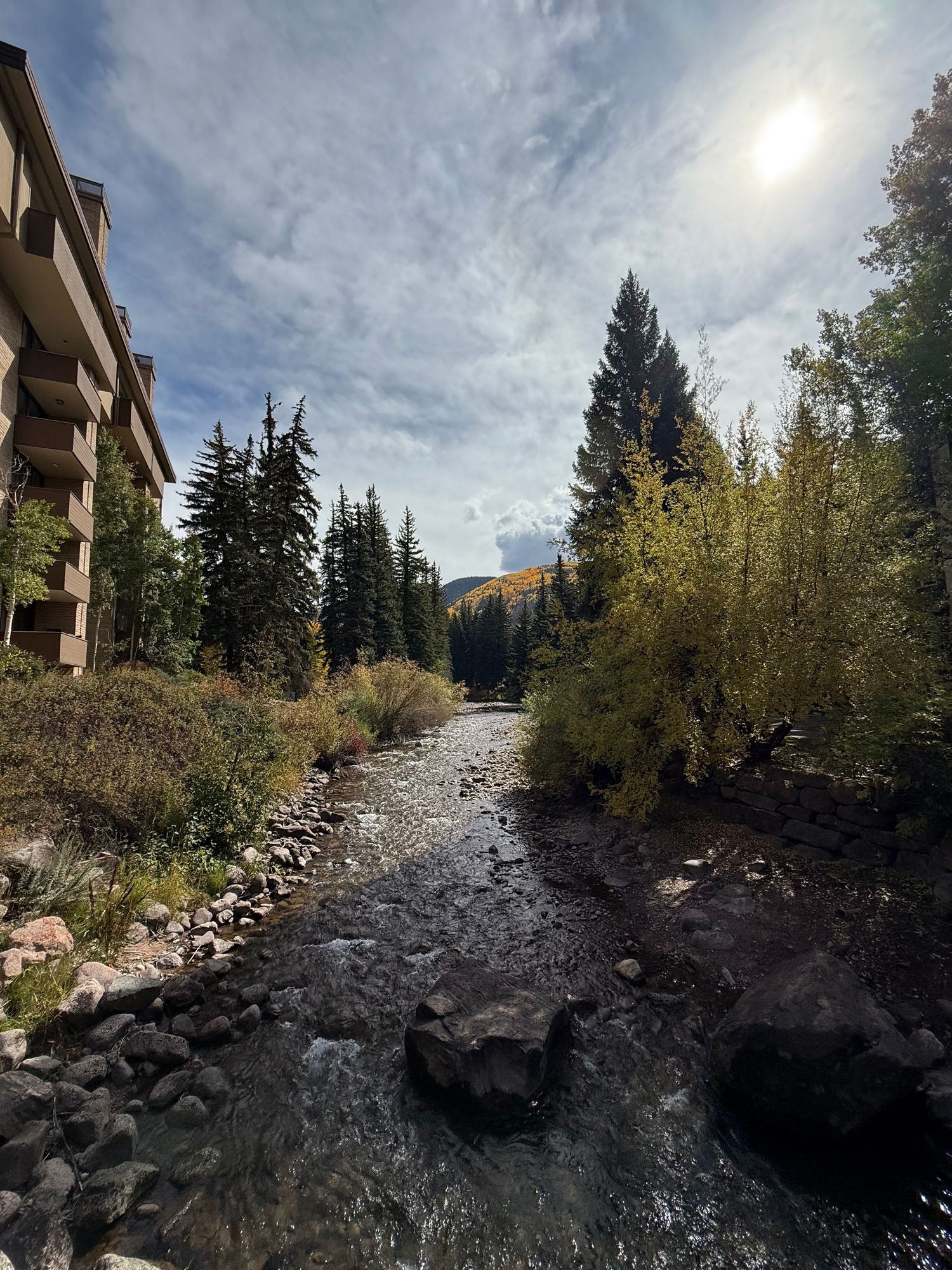 A rocky mountain stream flows past condominiums and evergreen trees under a bright cloudy sky in a scenic landscape.