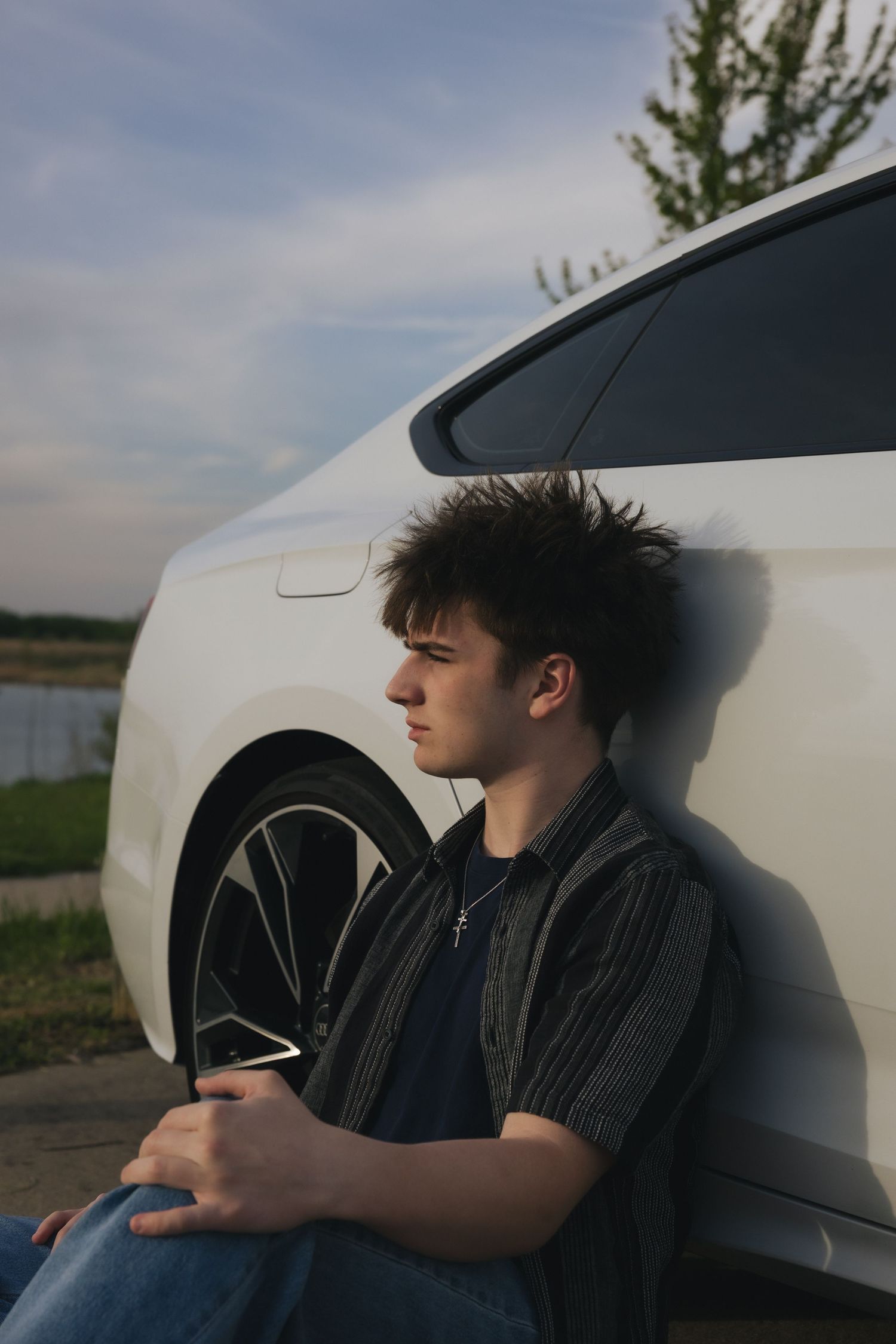 Young man with messy hair leaning against a white car outdoors, holding a phone, looking thoughtful.