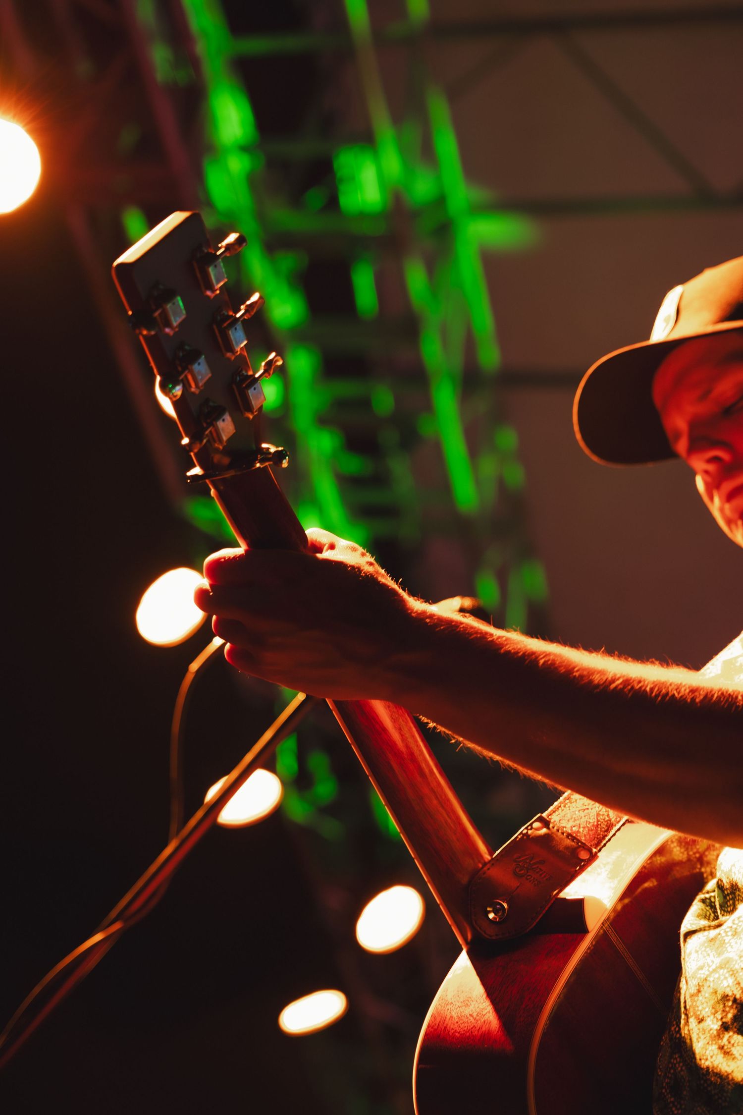 Musician in a hat plays acoustic guitar on stage under green and warm lights during a live concert.